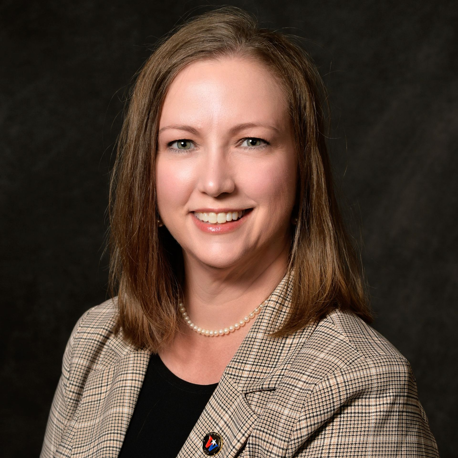 Woman in a blazer and pearl necklace smiles, headshot against a dark background.