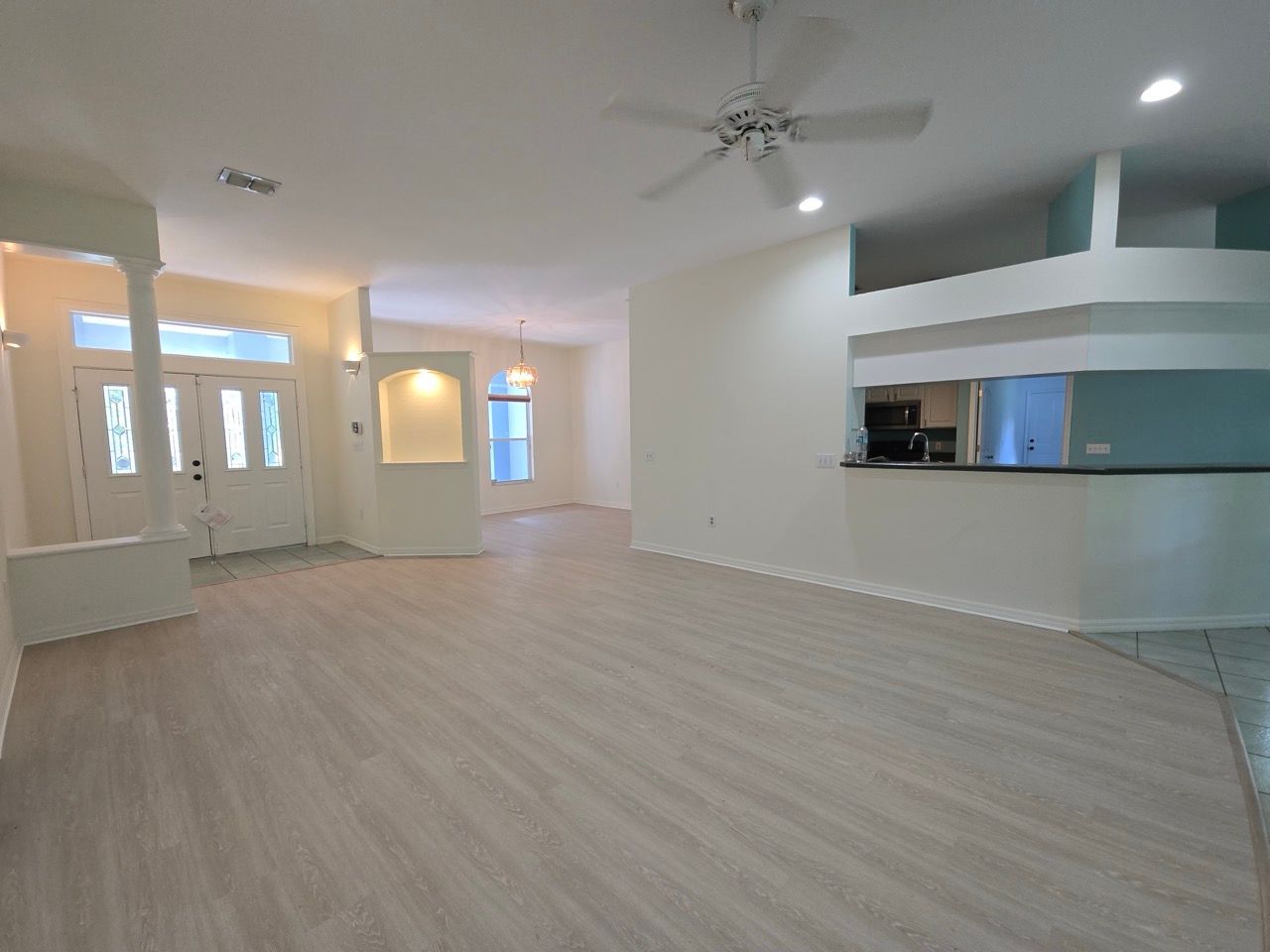Empty modern living room with light wood floors, white walls, ceiling fan, and open kitchen pass-through.