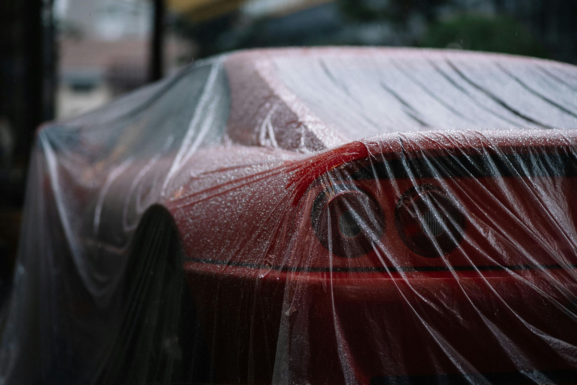 Red car covered in a clear tarp, beaded with water droplets, parked outdoors.