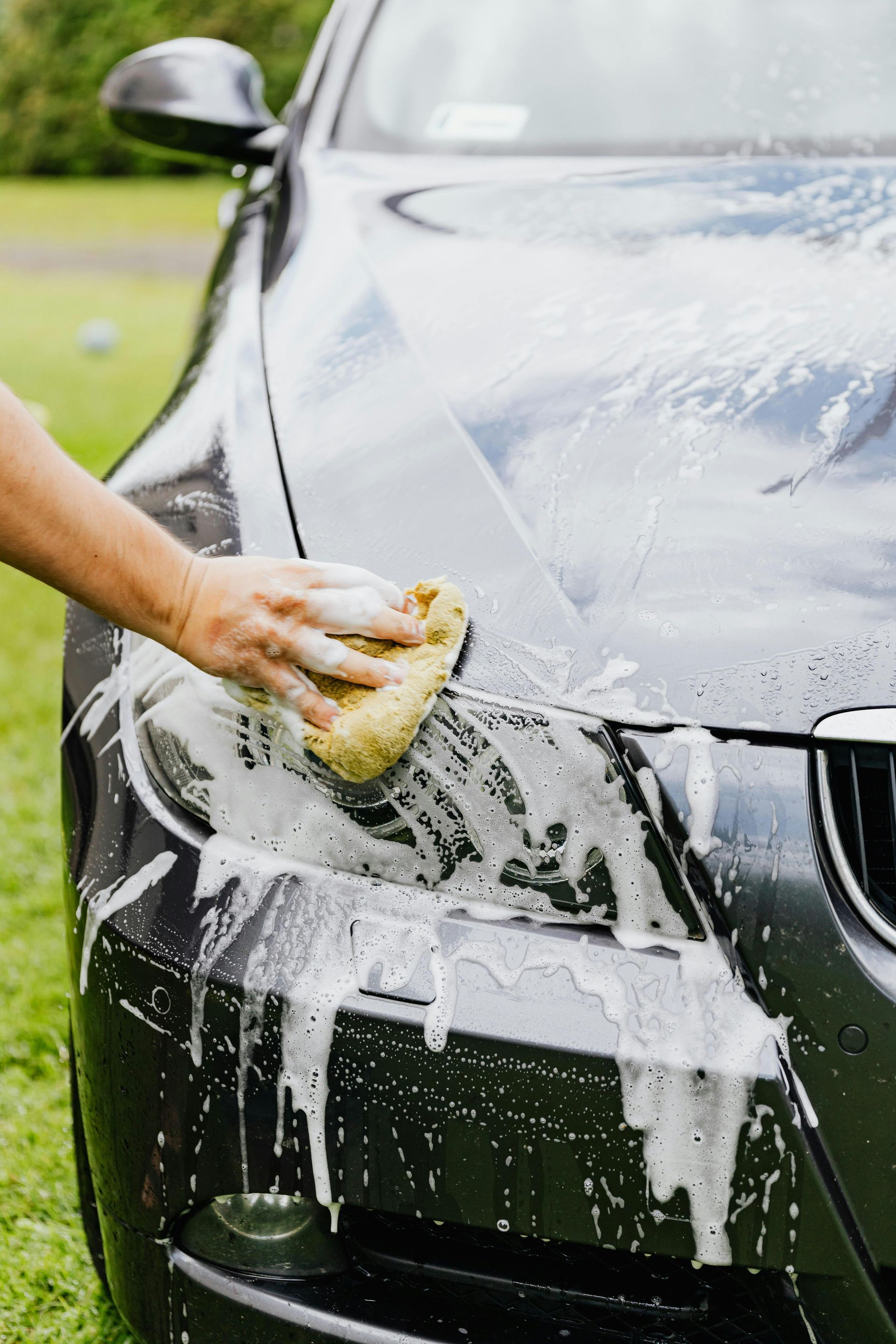 Person washing a dark gray car with a soapy sponge outdoors on the grass.