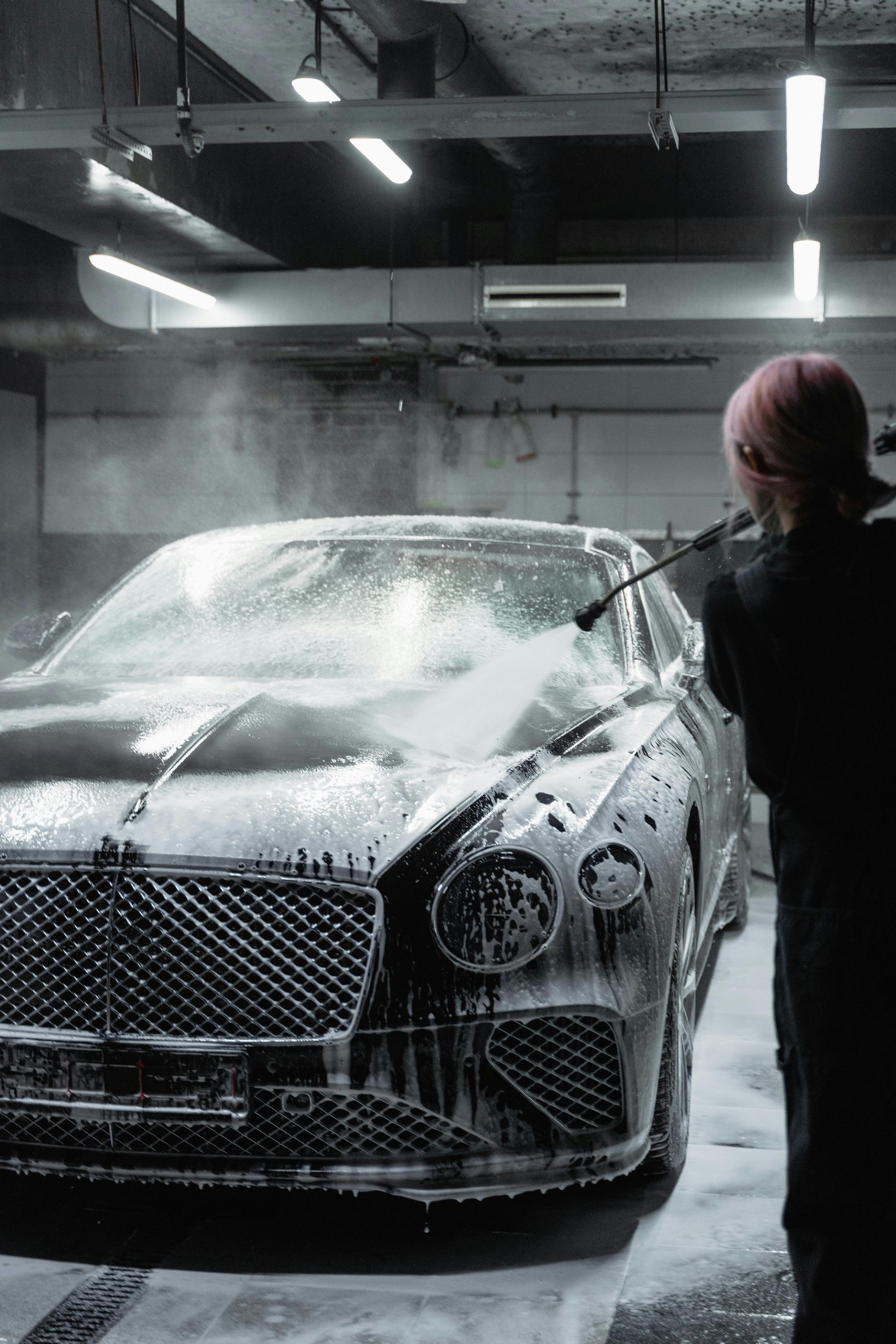 Person washing a black Bentley with a pressure washer in a garage.