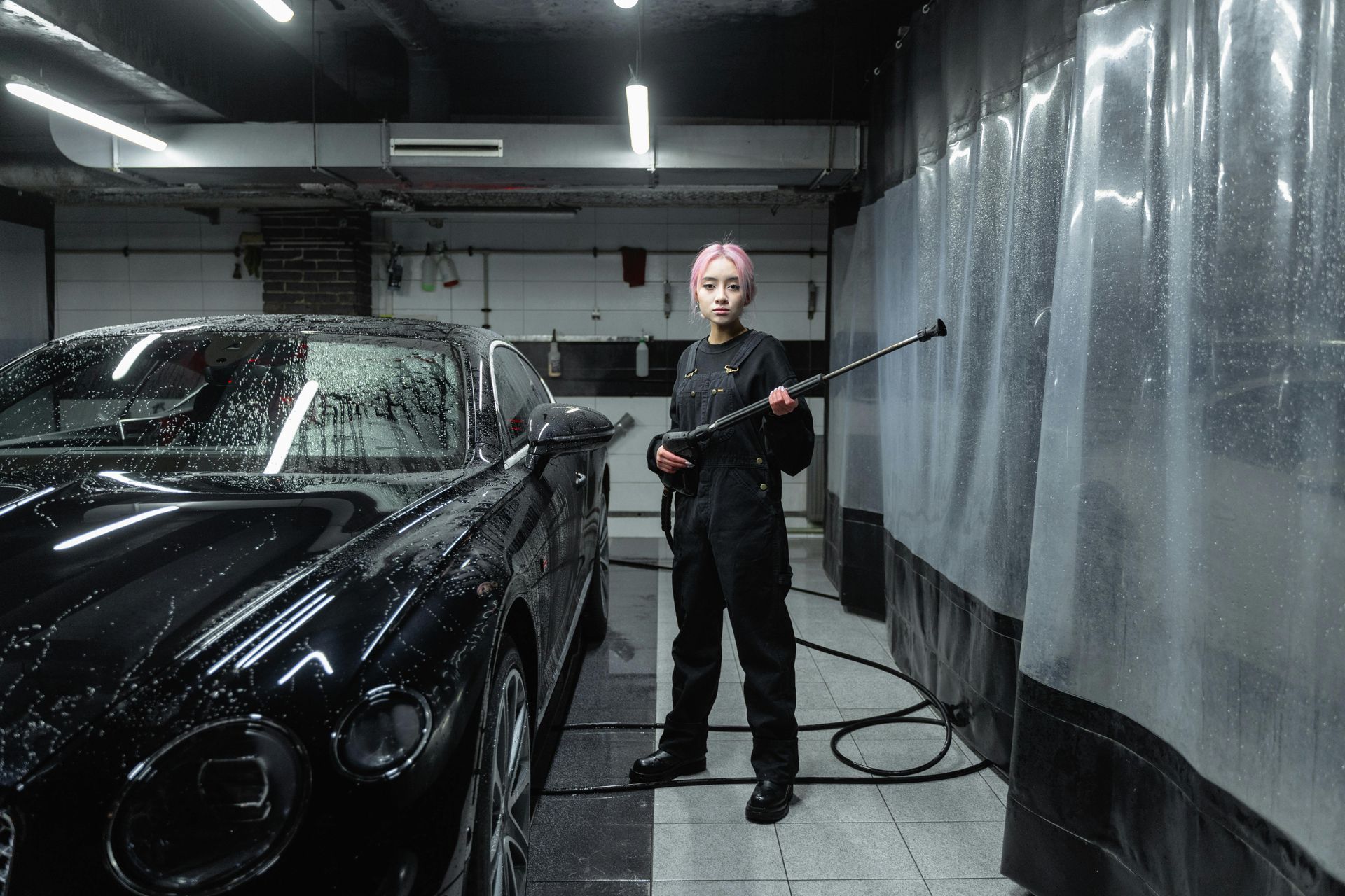 Person with pink hair washes a black car in a car wash, holding a spray nozzle.