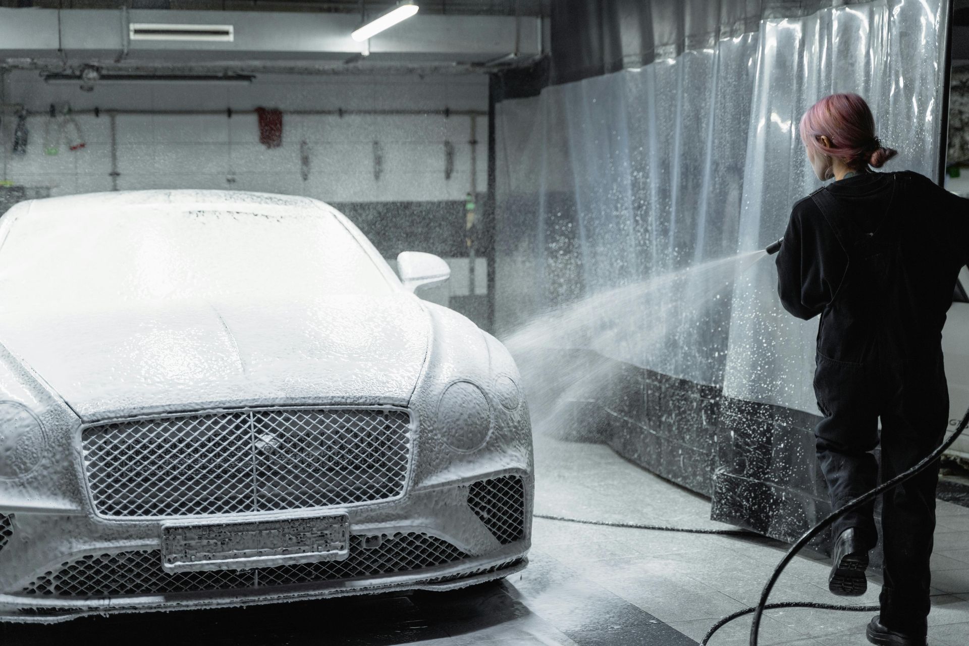 Person washing a car with foamy soap. The car is silver and in a car wash.