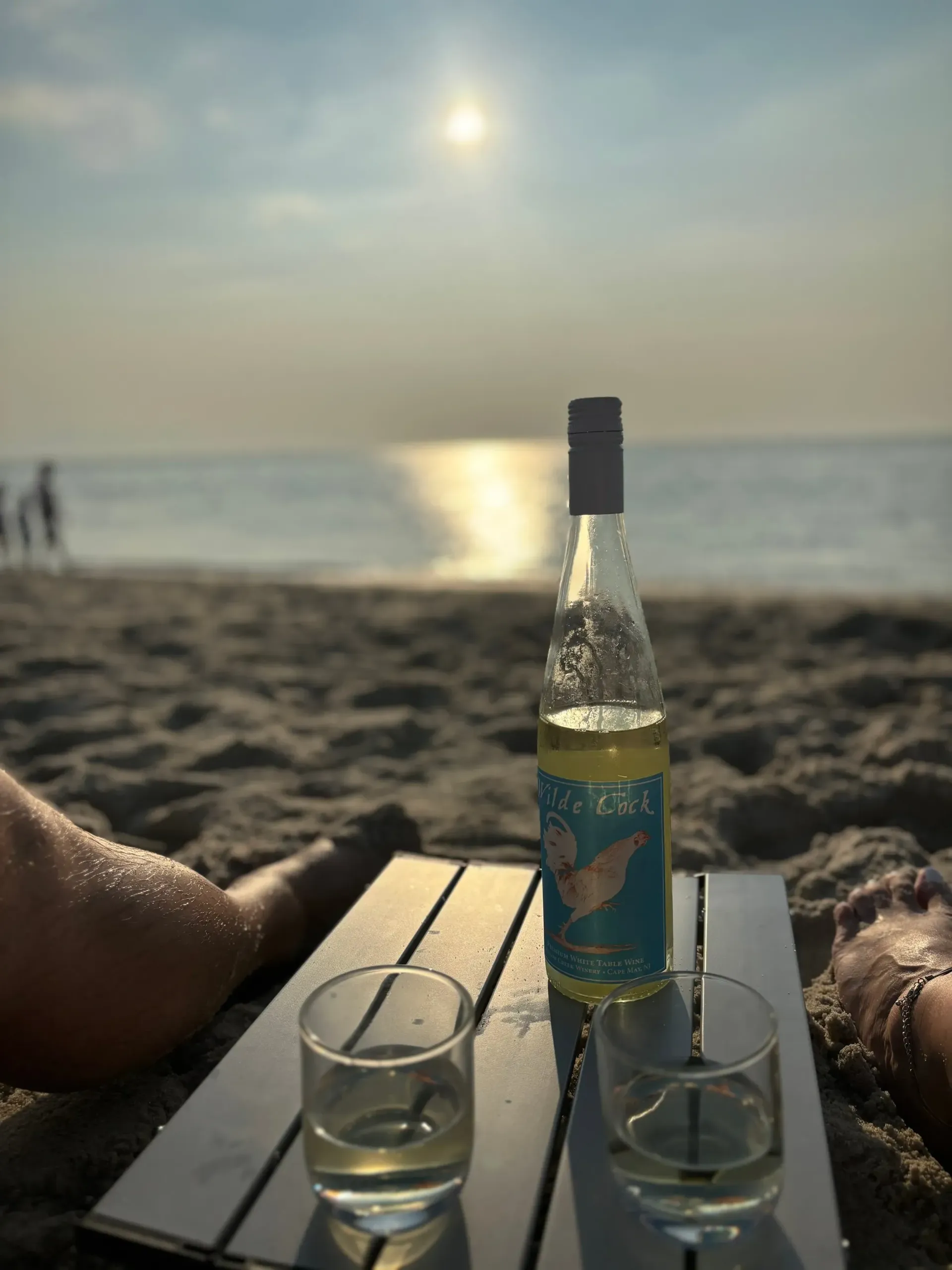A person is laying on the beach with a bottle and two glasses