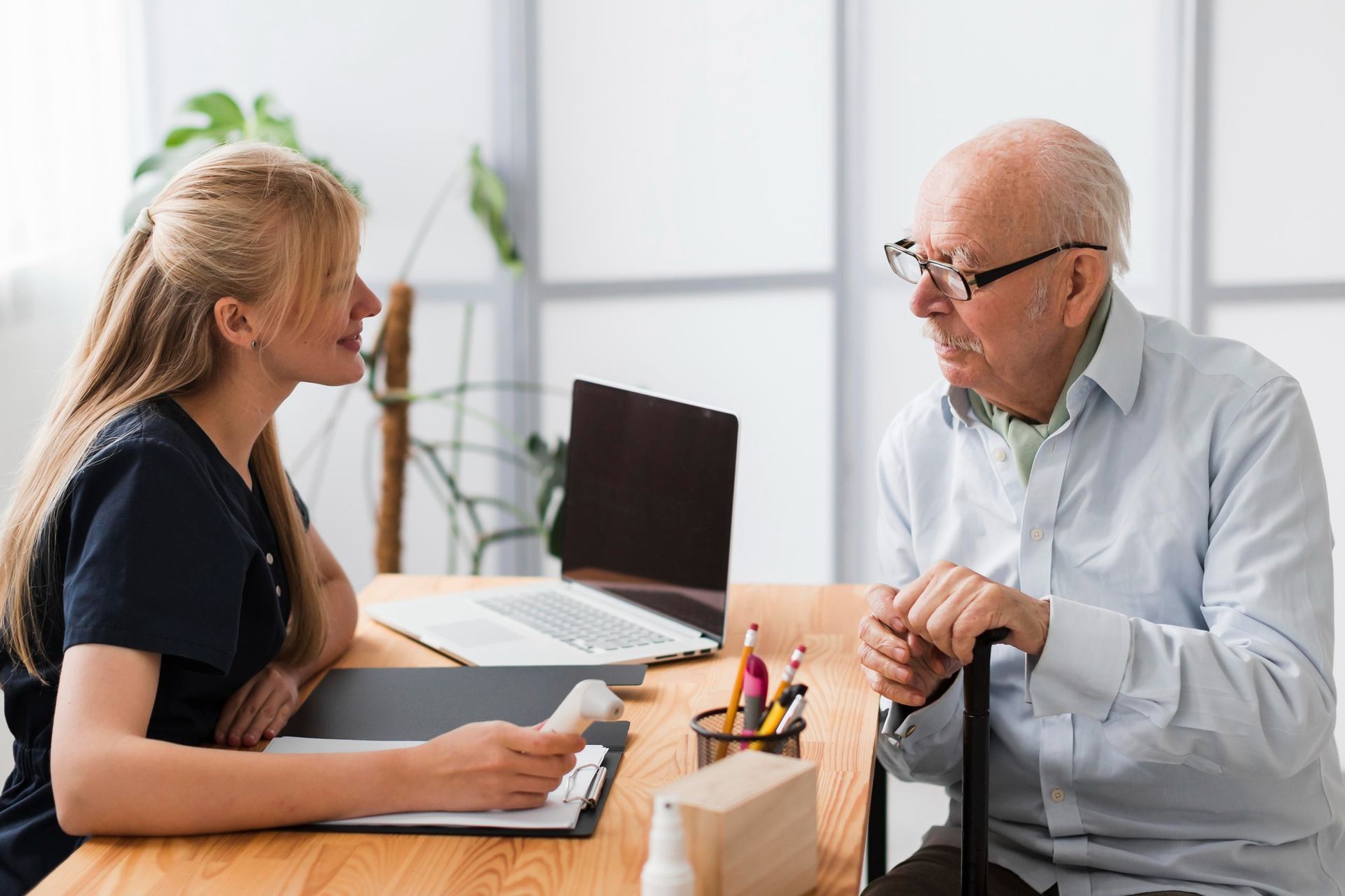 Young woman in scrubs consults with elderly man, holding prescription. They are seated at a desk, laptop on desk.