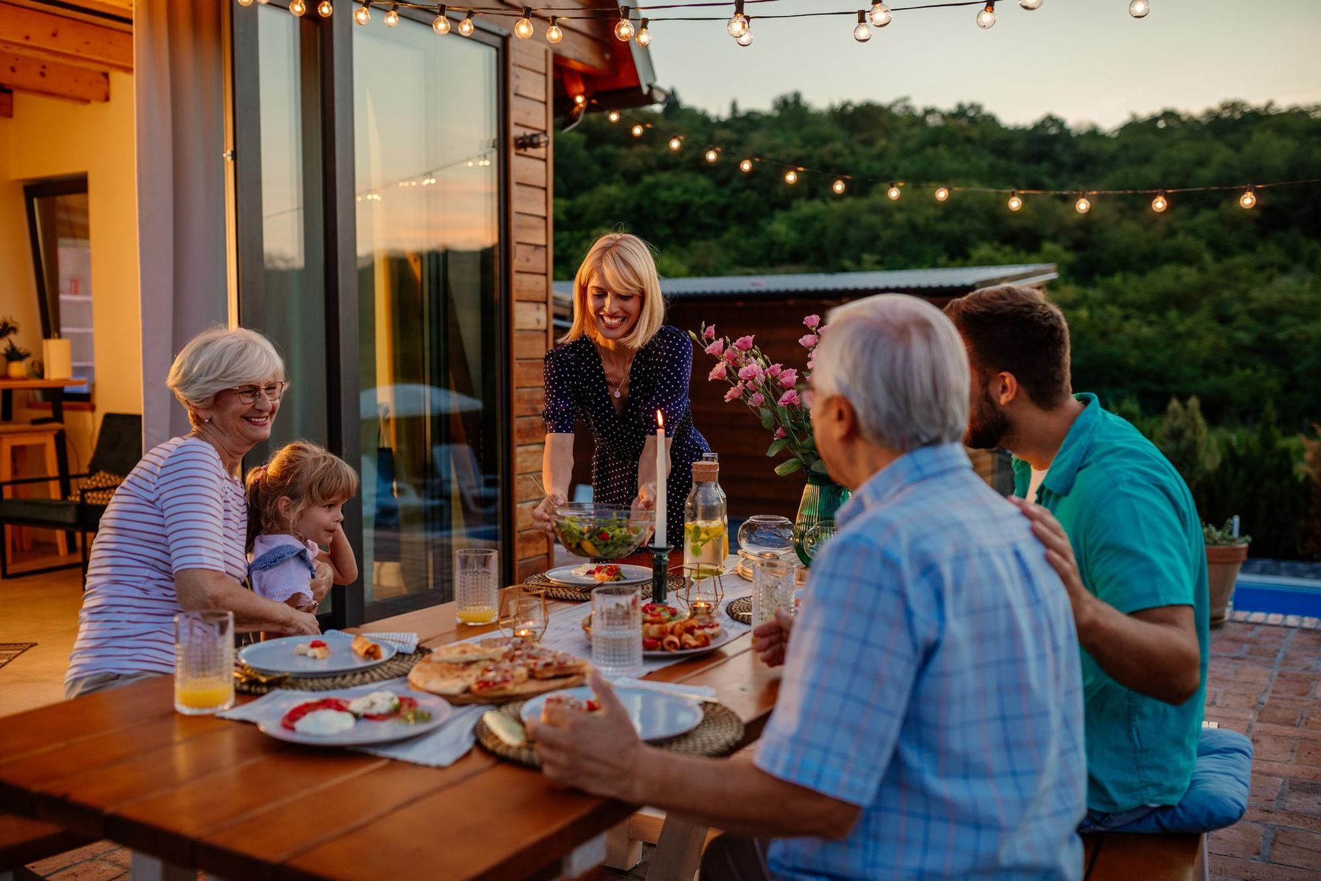Family gathering outdoors around a table. Woman serves salad. String lights illuminate the evening.