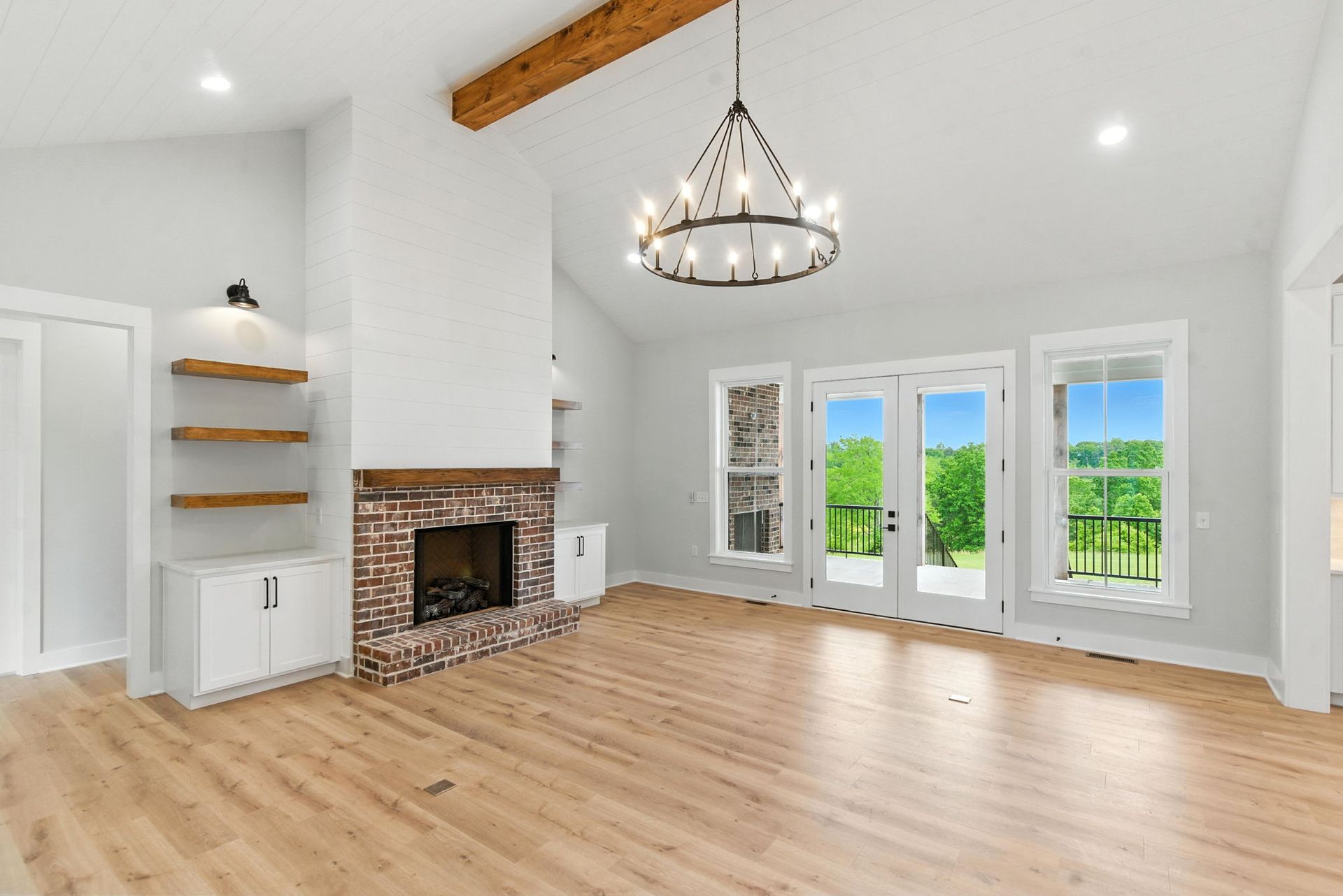 Bright empty living room with hardwood floors, brick fireplace, chandelier, and large windows overlooking greenery