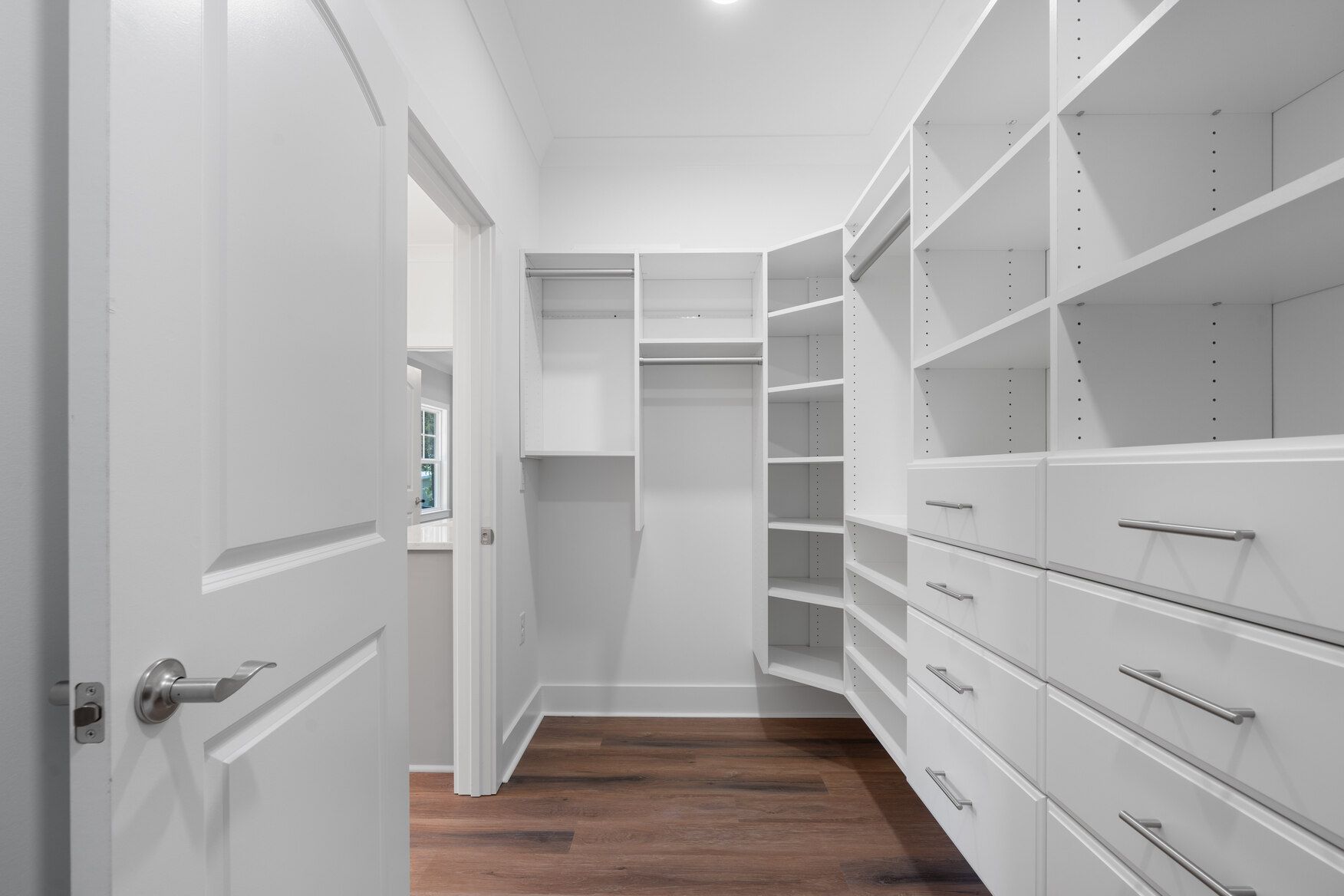 Bright white walk-in closet with open shelving, drawers, and wood flooring, viewed through a doorway