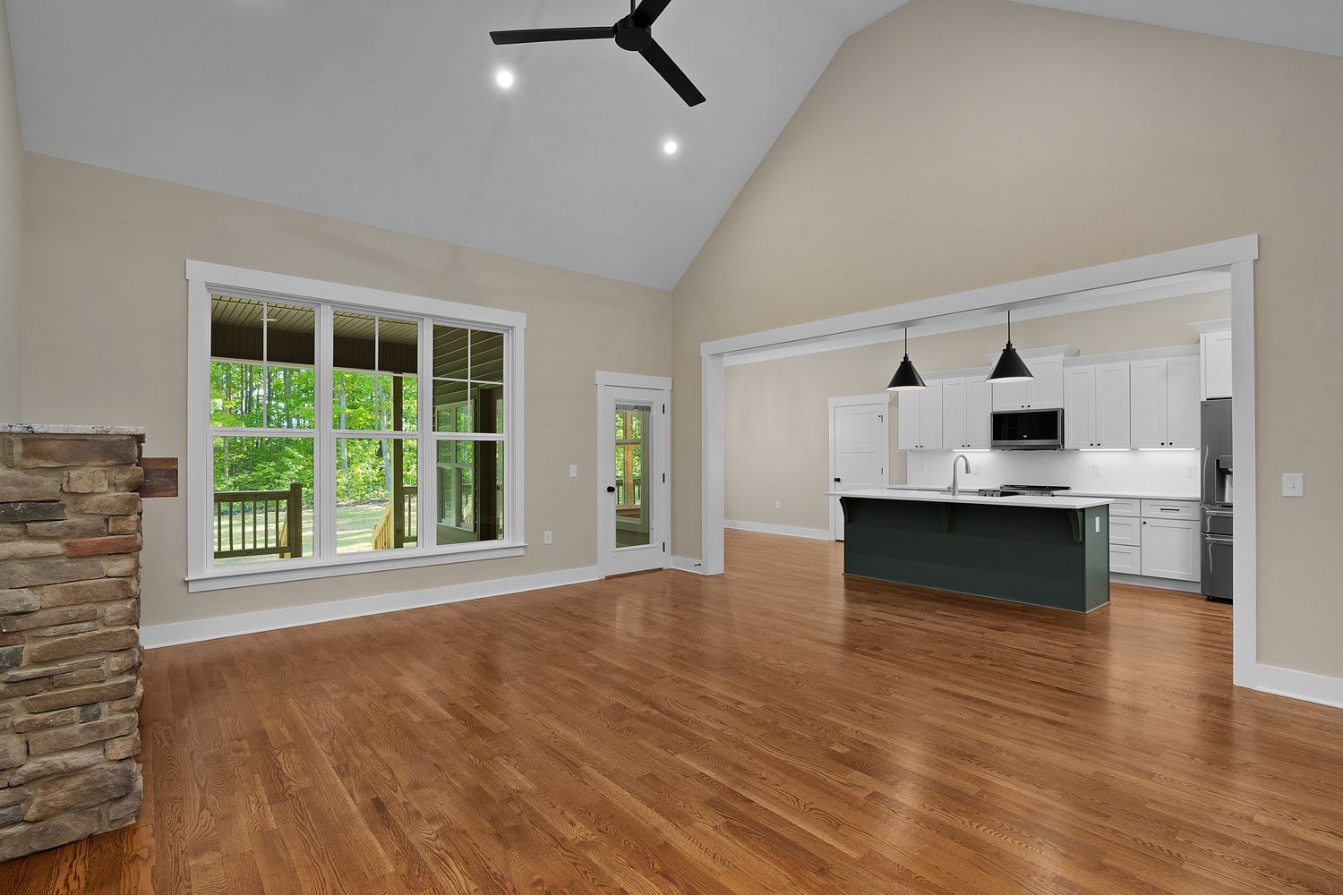 Spacious empty living room with vaulted ceiling, large window, stone fireplace, and open kitchen view