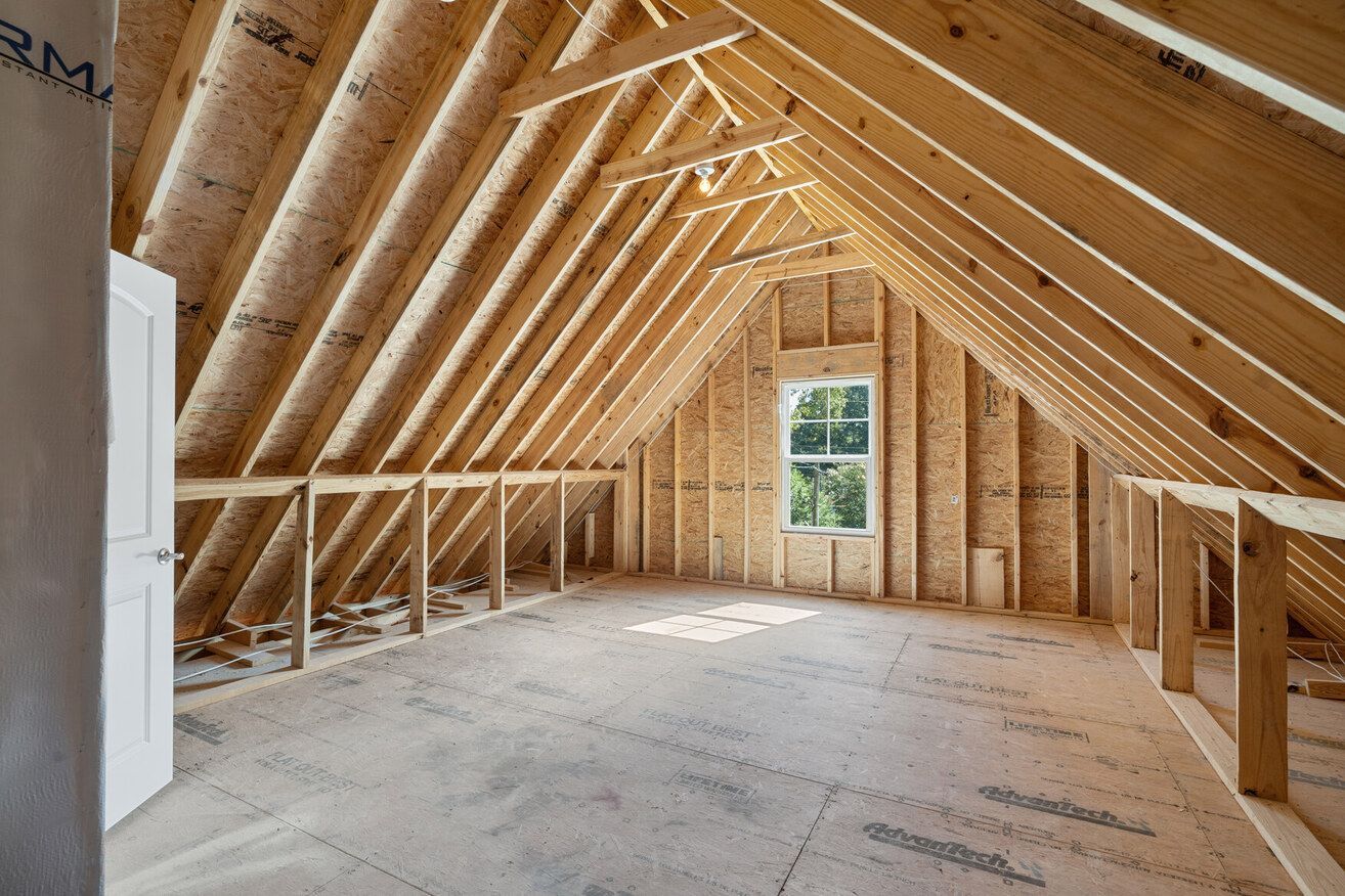 Unfinished attic with exposed wooden rafters, insulation, and a small window at the far end.