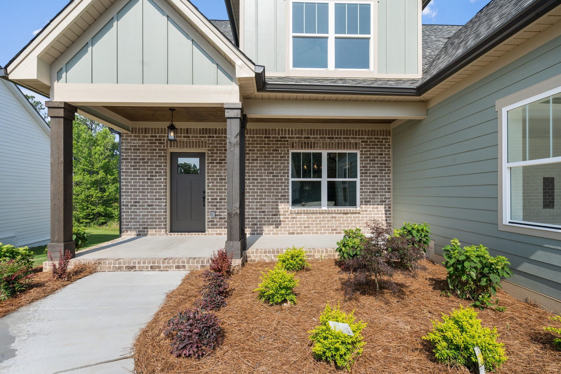 Front entrance of a brick house with covered porch, walkway, and landscaped mulch beds with small shrubs.