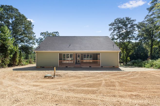 Front view of a small beige house with a gray roof, front porch, and gravel yard surrounded by trees
