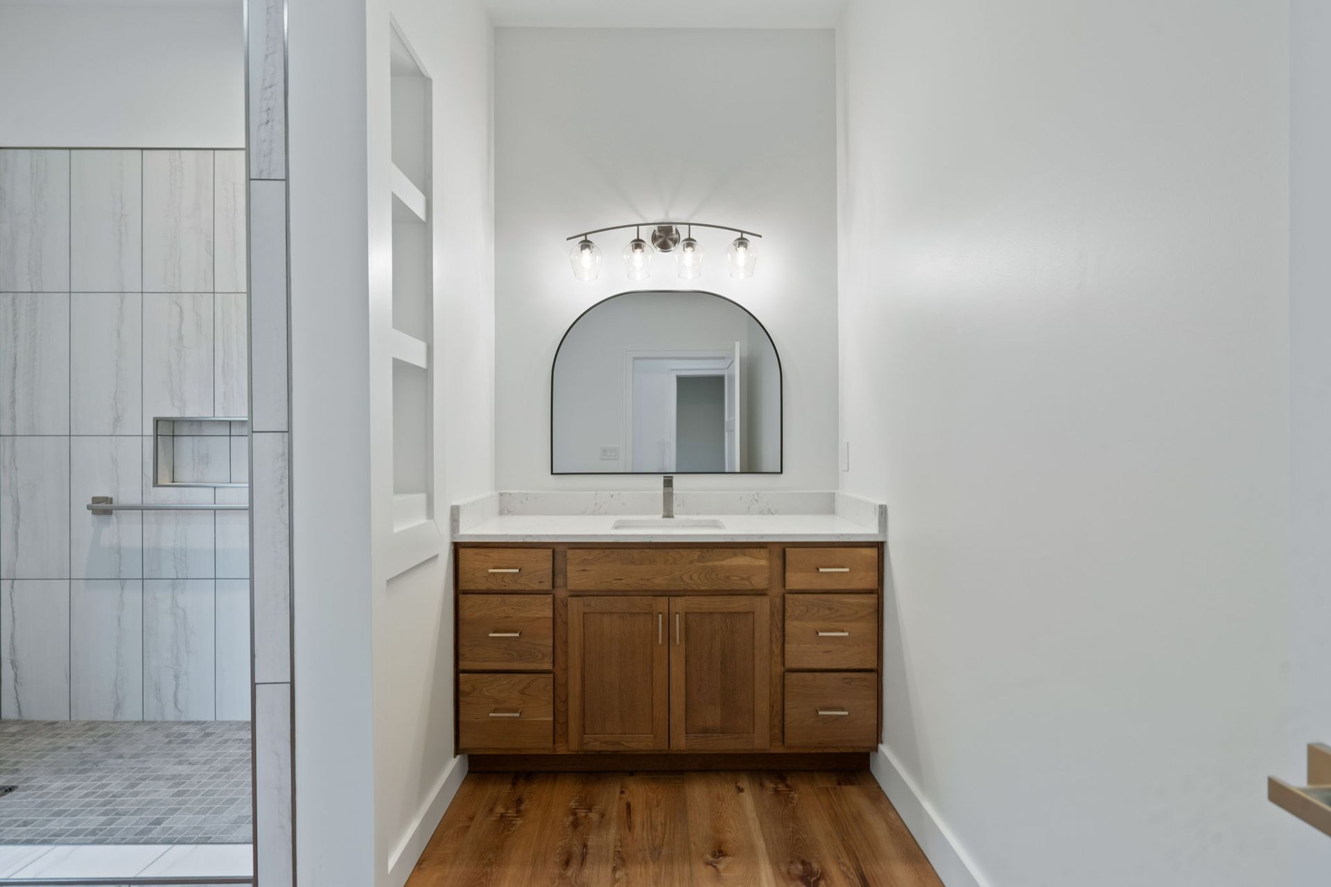 Bathroom vanity with wood cabinets, arched mirror, and white walls beside a tiled shower.