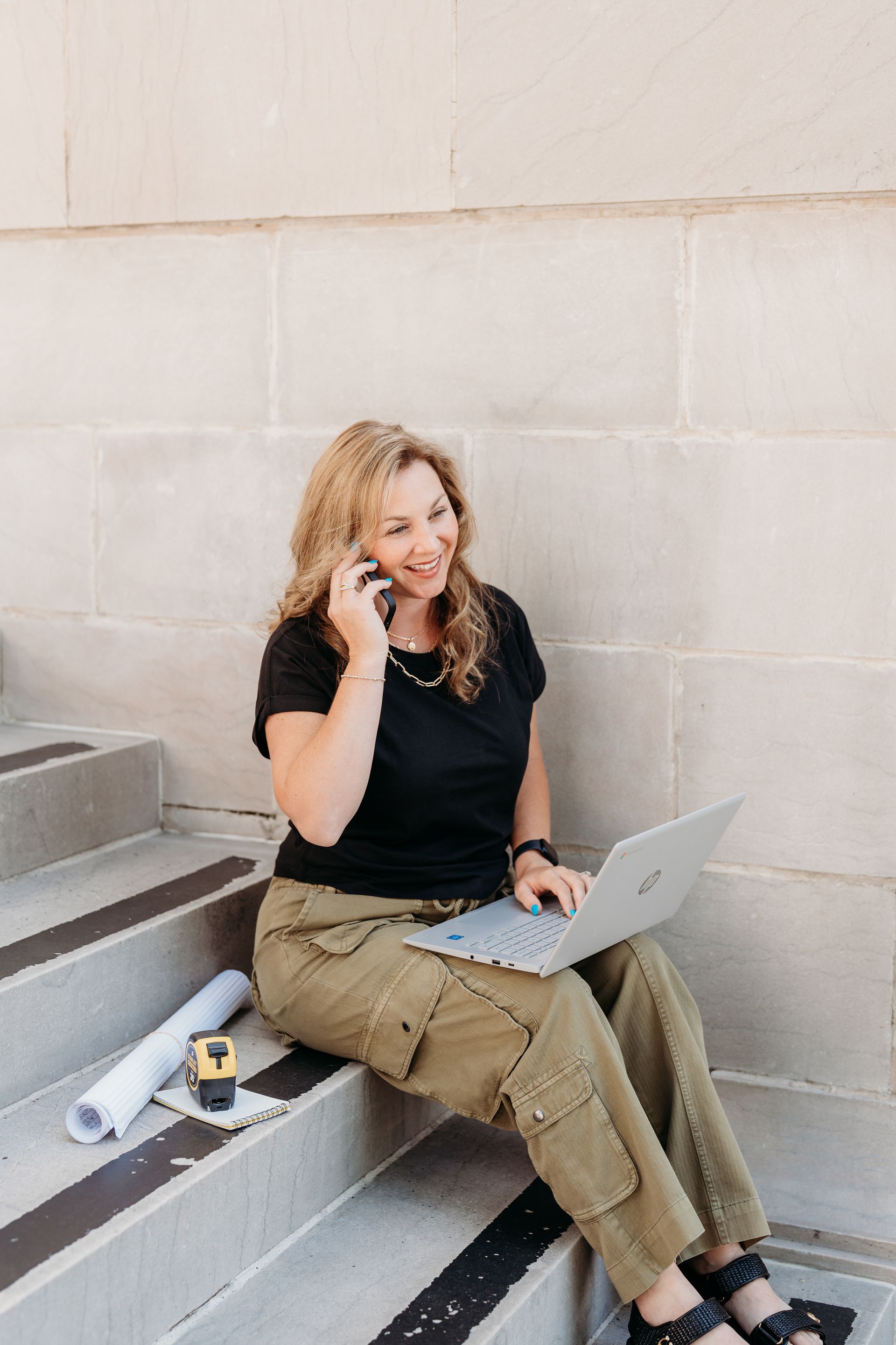 Woman sitting on steps with a laptop and phone, smiling against a light stone wall