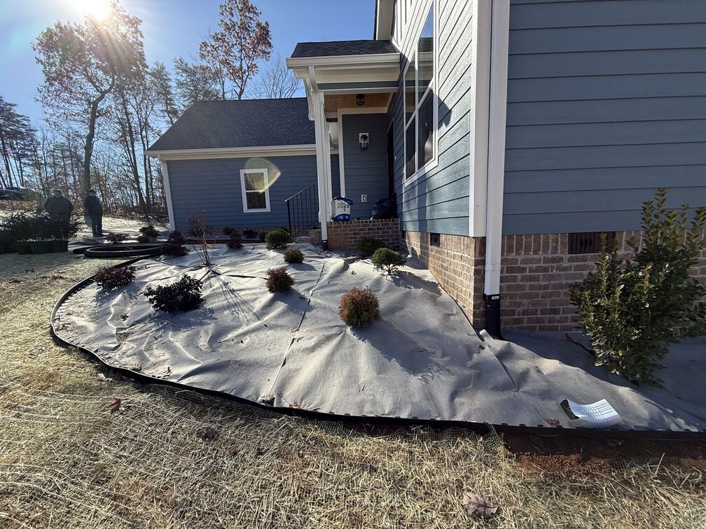 Front yard landscaping with gray weed barrier fabric and small shrubs beside a house in bright sunlight