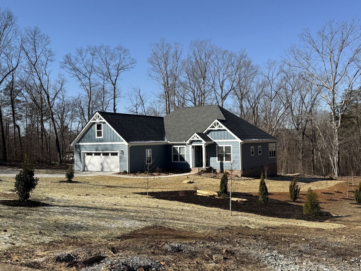 Small gray house with white trim and attached garage on a wooded lot under a clear blue sky