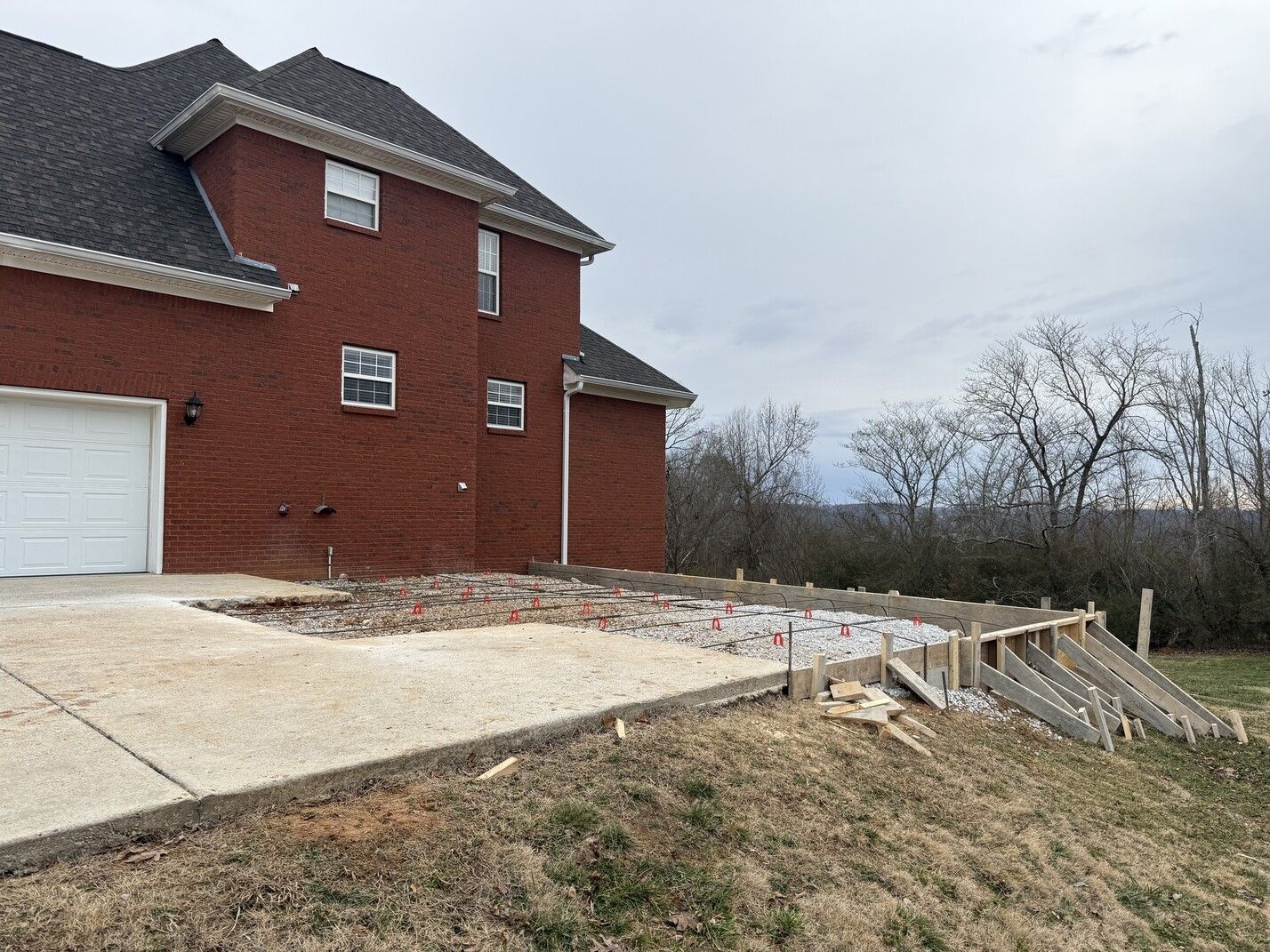 Brick house with a garage and cracked concrete driveway beside a sloped yard and trees