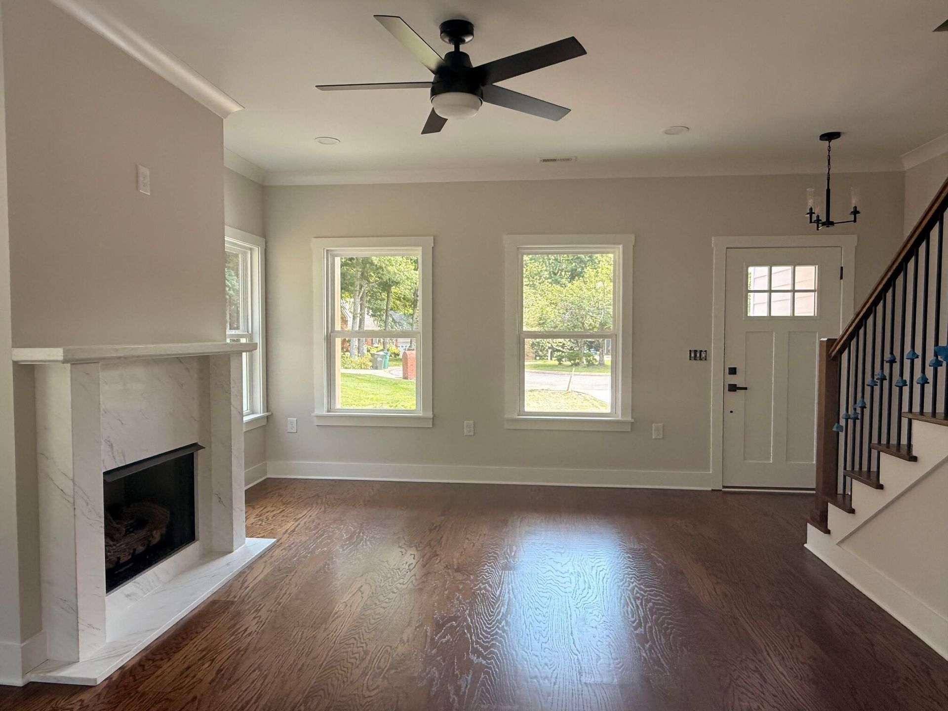Empty living room with fireplace, hardwood floors, ceiling fan, two windows, and staircase.