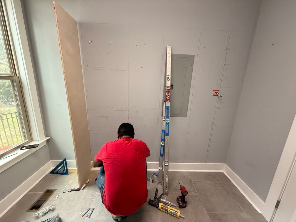 Worker kneeling on a floor in a room under renovation, with tools and a ladder against the wall.