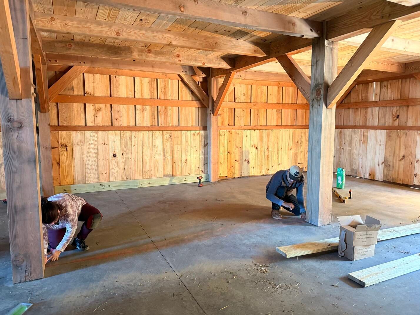 Two workers crouch in a wooden barn interior with exposed beams and concrete floor during construction.