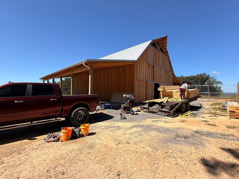 Rustic wooden barn under construction beside a red pickup, with lumber, tools, and dirt yard.