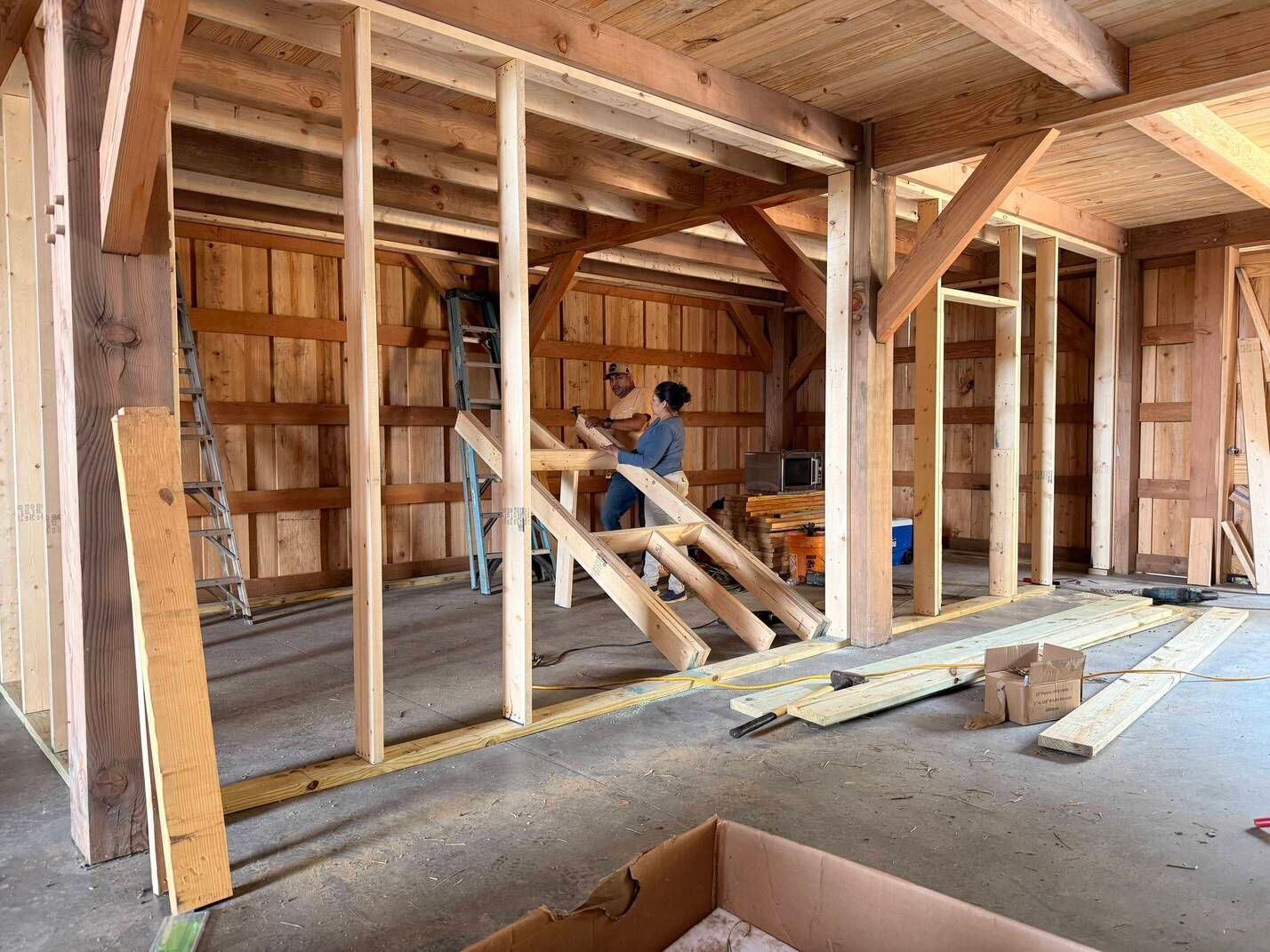 Wooden barn interior under renovation, with framing beams, tools, and a worker on a ladder.