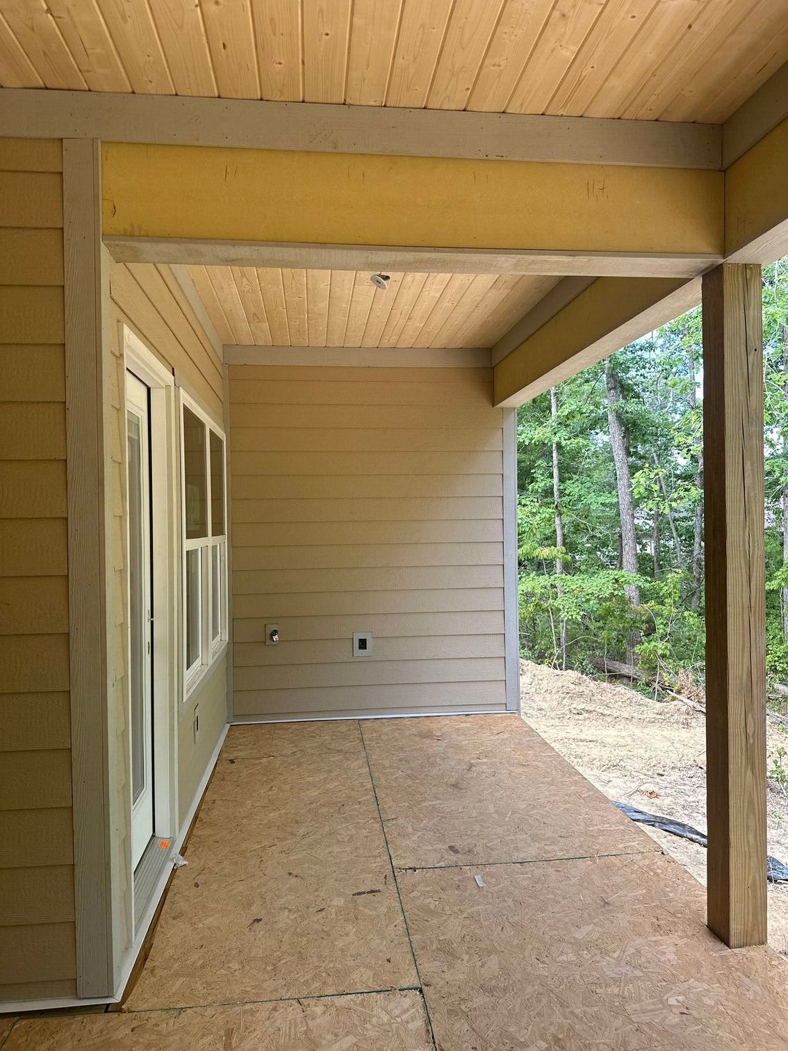 Covered porch with wood ceiling, screened window, and unfinished floor overlooking trees.
