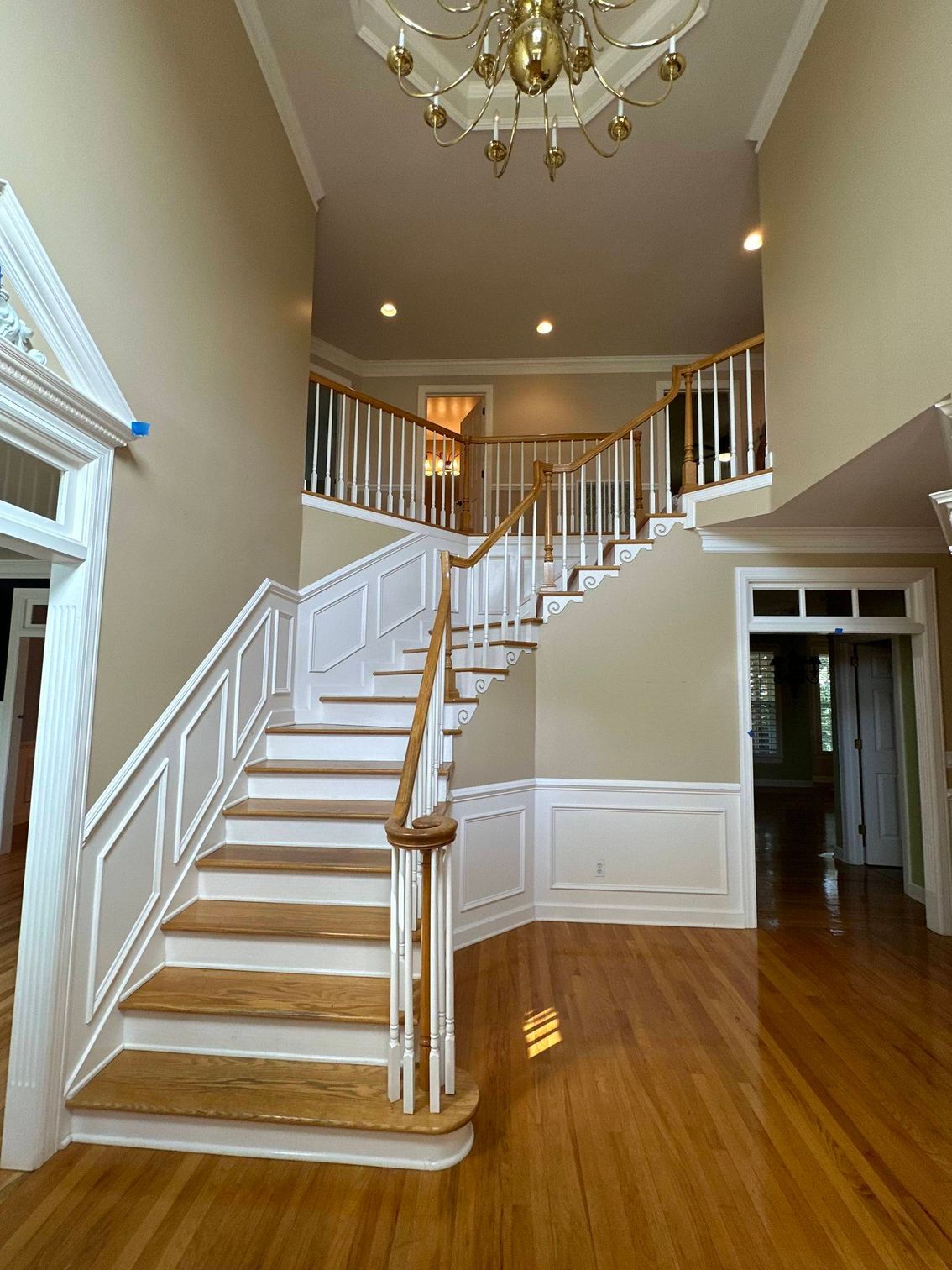 Bright foyer with white staircase, wood floors, chandelier, and upper balcony railing.