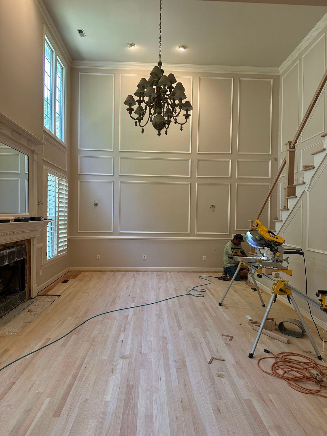 Empty living room with light wood floors, white paneled walls, chandelier, and tools near a staircase during renovation
