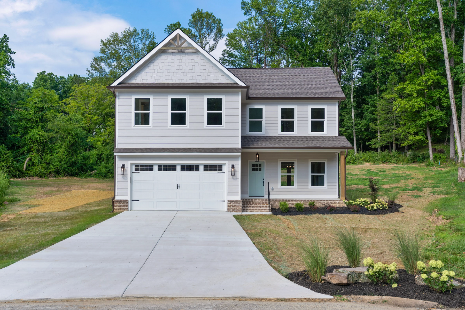 Two-story gray suburban house with a white garage, concrete driveway, and green wooded yard