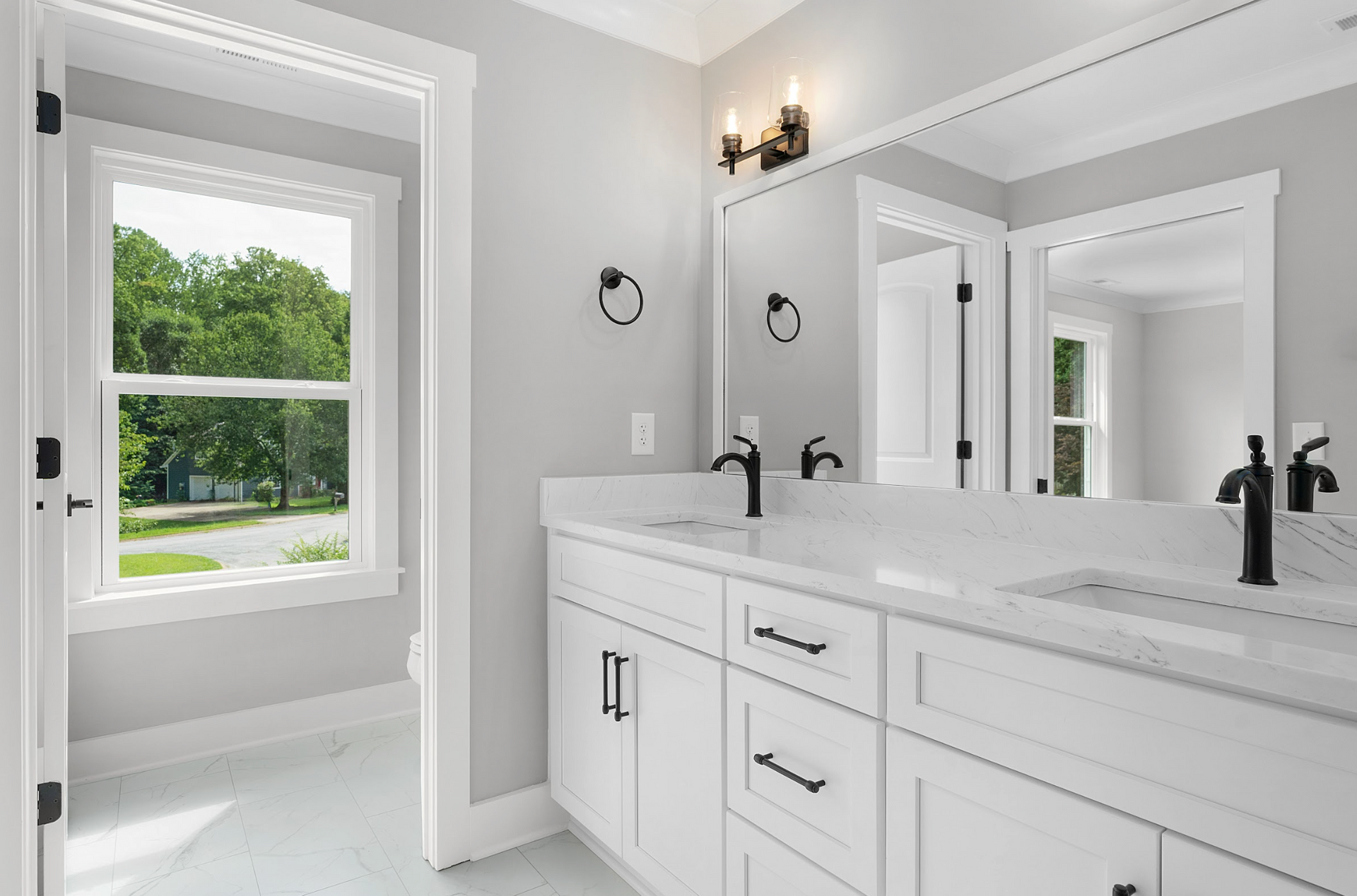 Bright white bathroom with double vanity, large mirror, and window overlooking greenery.