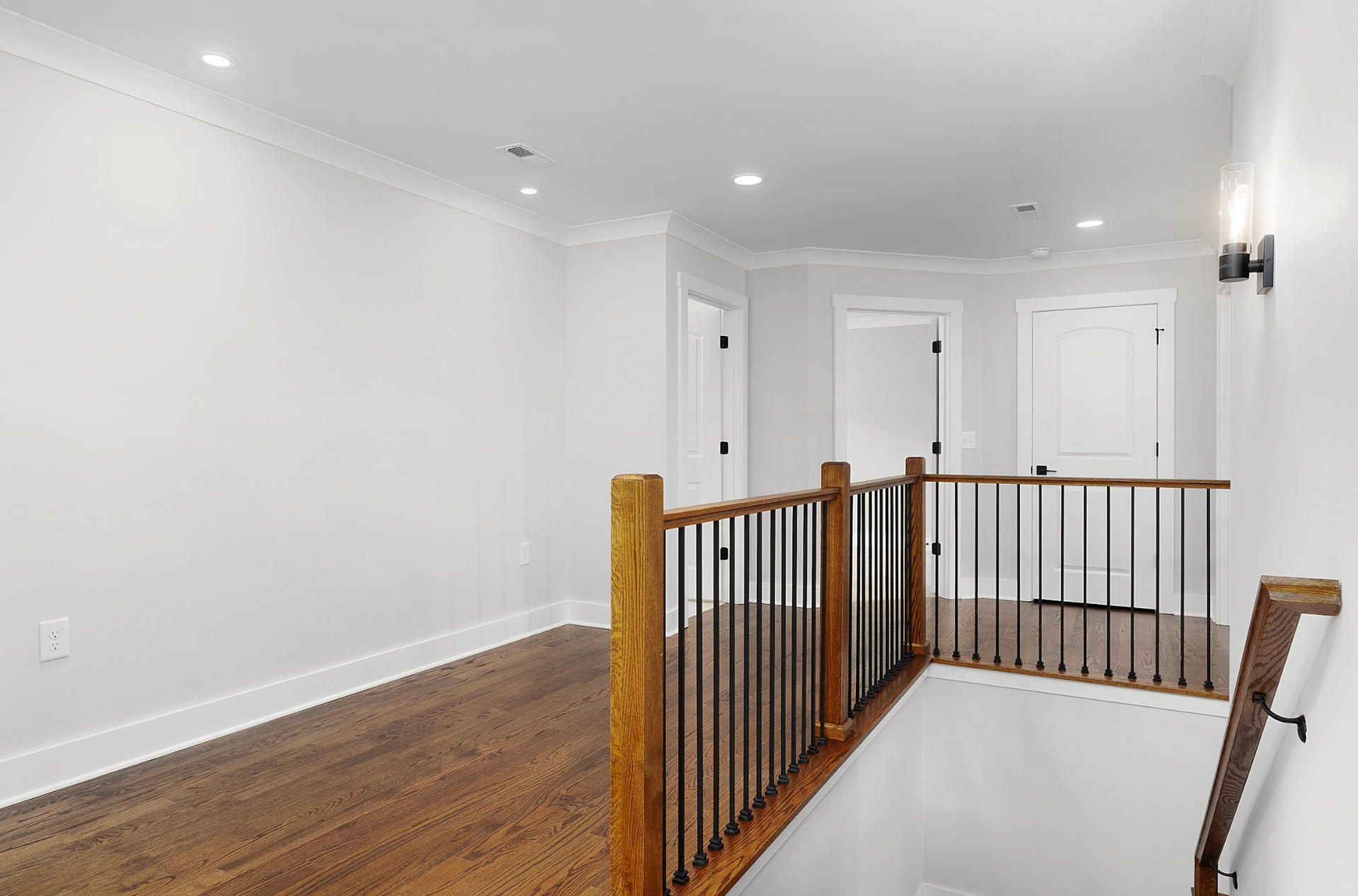 Bright hallway with white walls, hardwood floor, staircase railing, and closed doors.