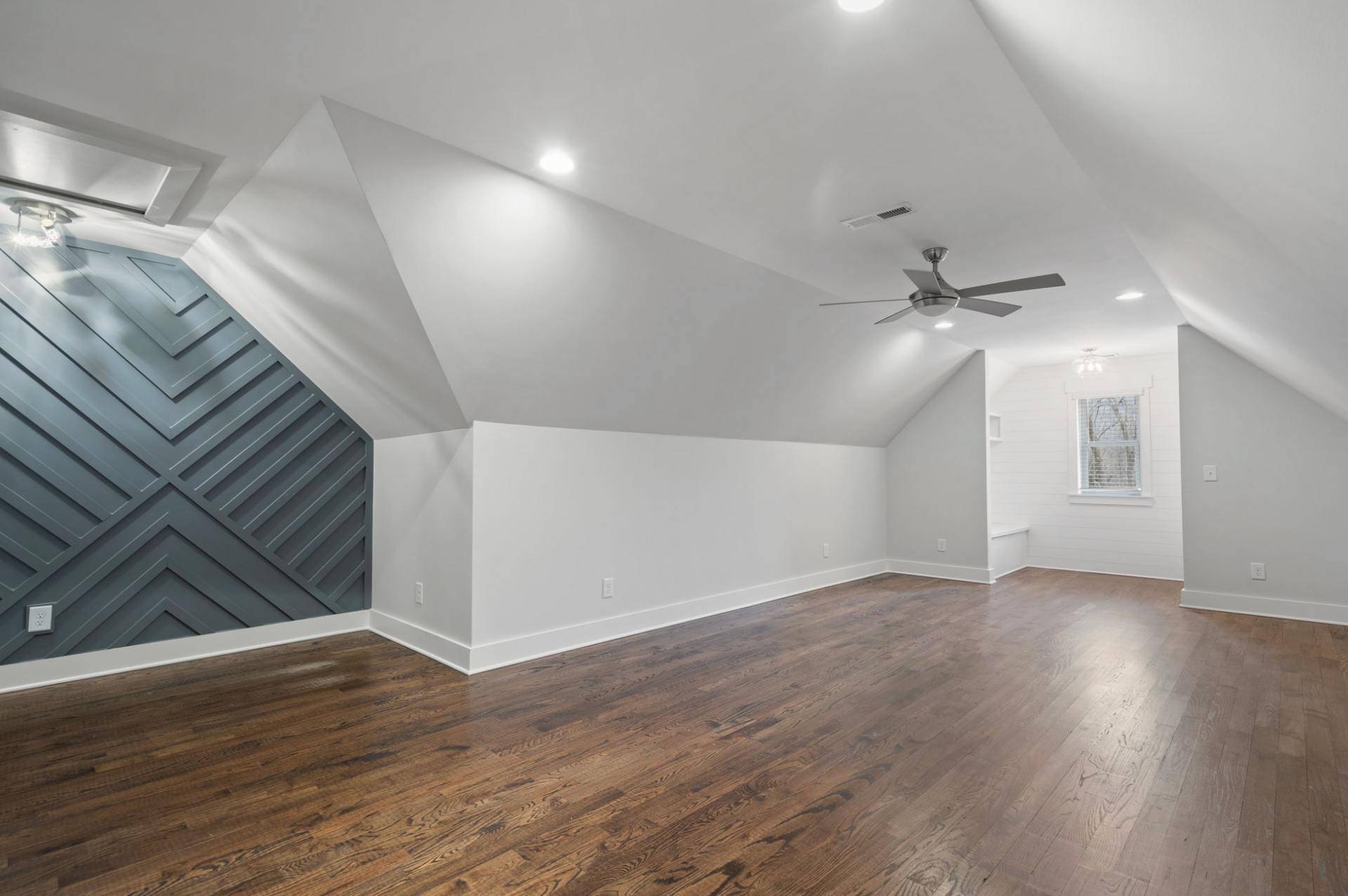 Empty modern attic room with sloped white ceiling, hardwood floors, ceiling fan, and blue accent wall