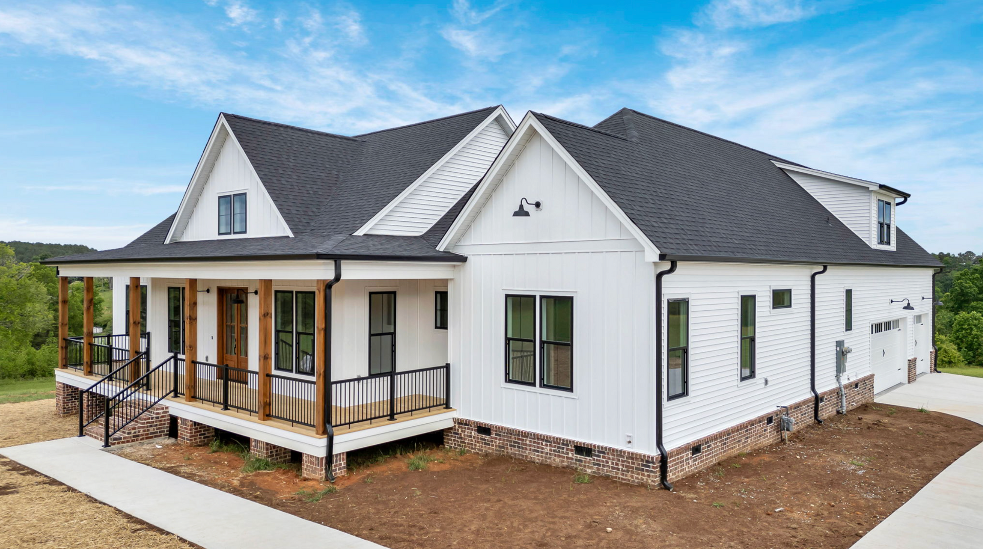White modern farmhouse with dark gray roof, wraparound porch, and brick foundation in a new residential area