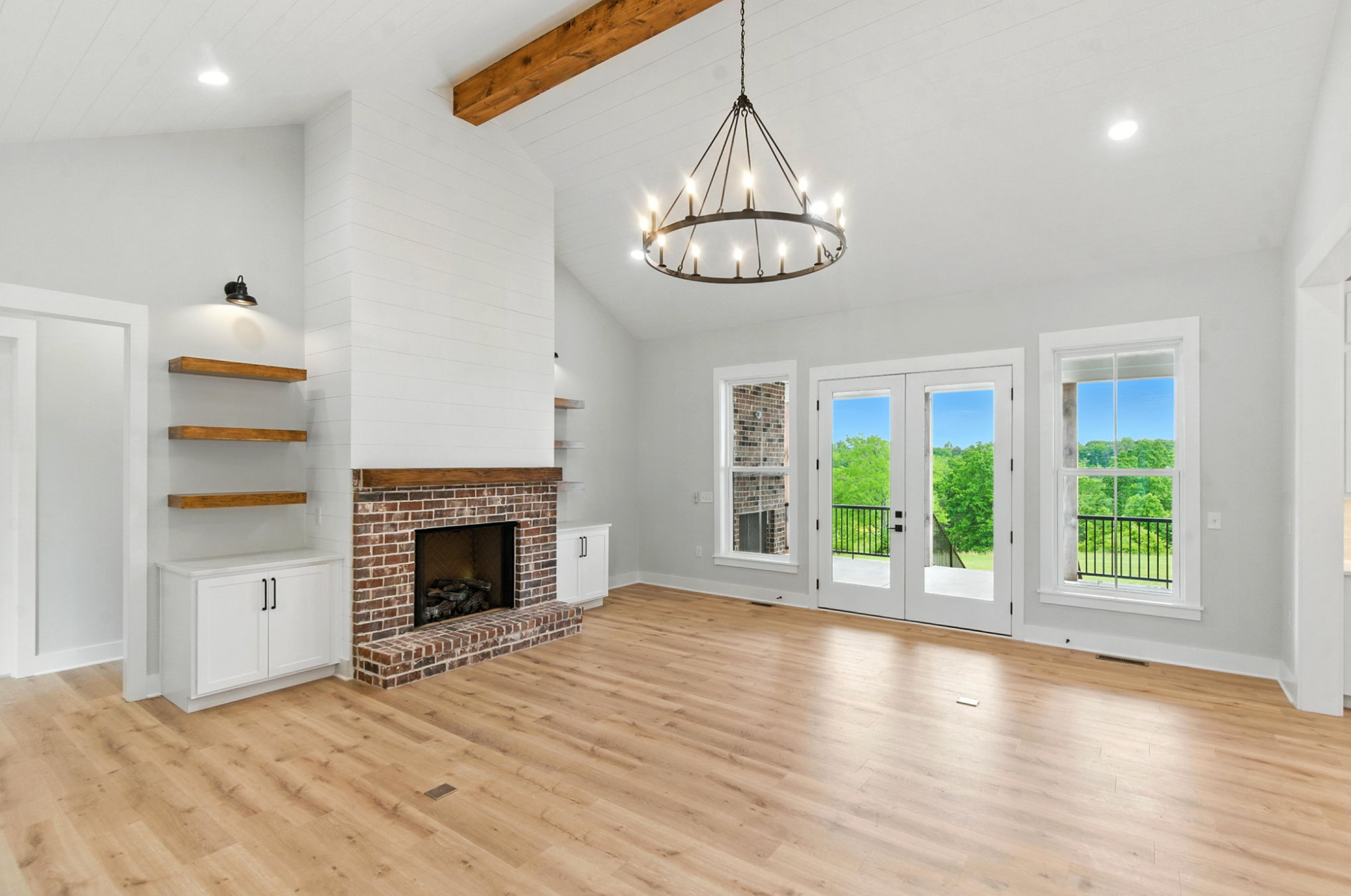 Bright open living room with wood floors, white walls, fireplace, chandelier, and large windows overlooking greenery