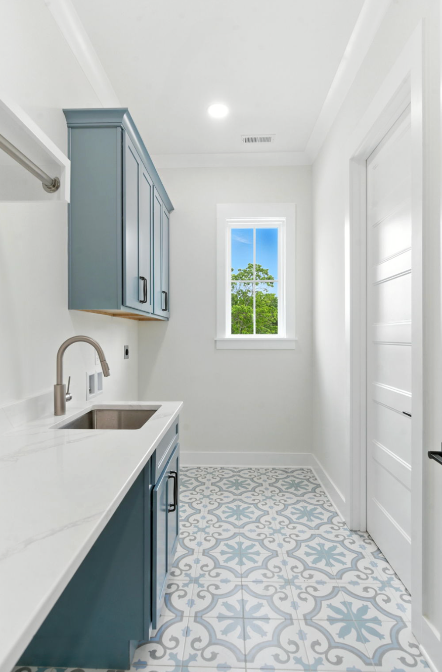 Bright laundry room with blue cabinets, white countertops, patterned tile floor, and a window.