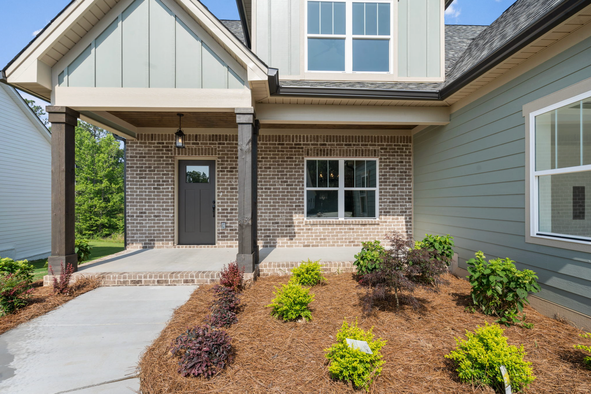 Front porch of a brick house with a covered entry, walkway, and landscaped mulch beds with shrubs.
