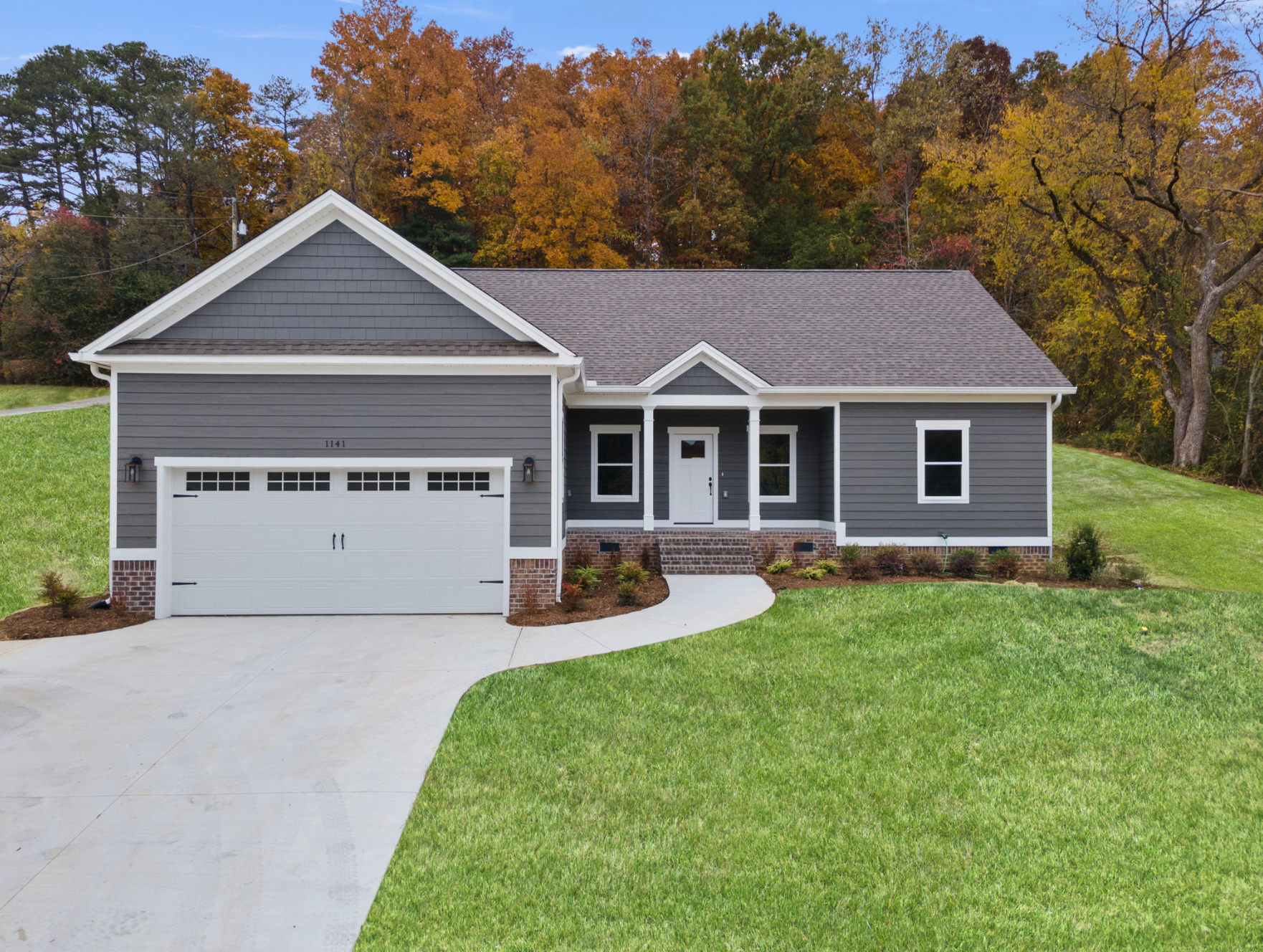 Gray single-story house with white garage, front porch, and curved driveway in a wooded suburban setting