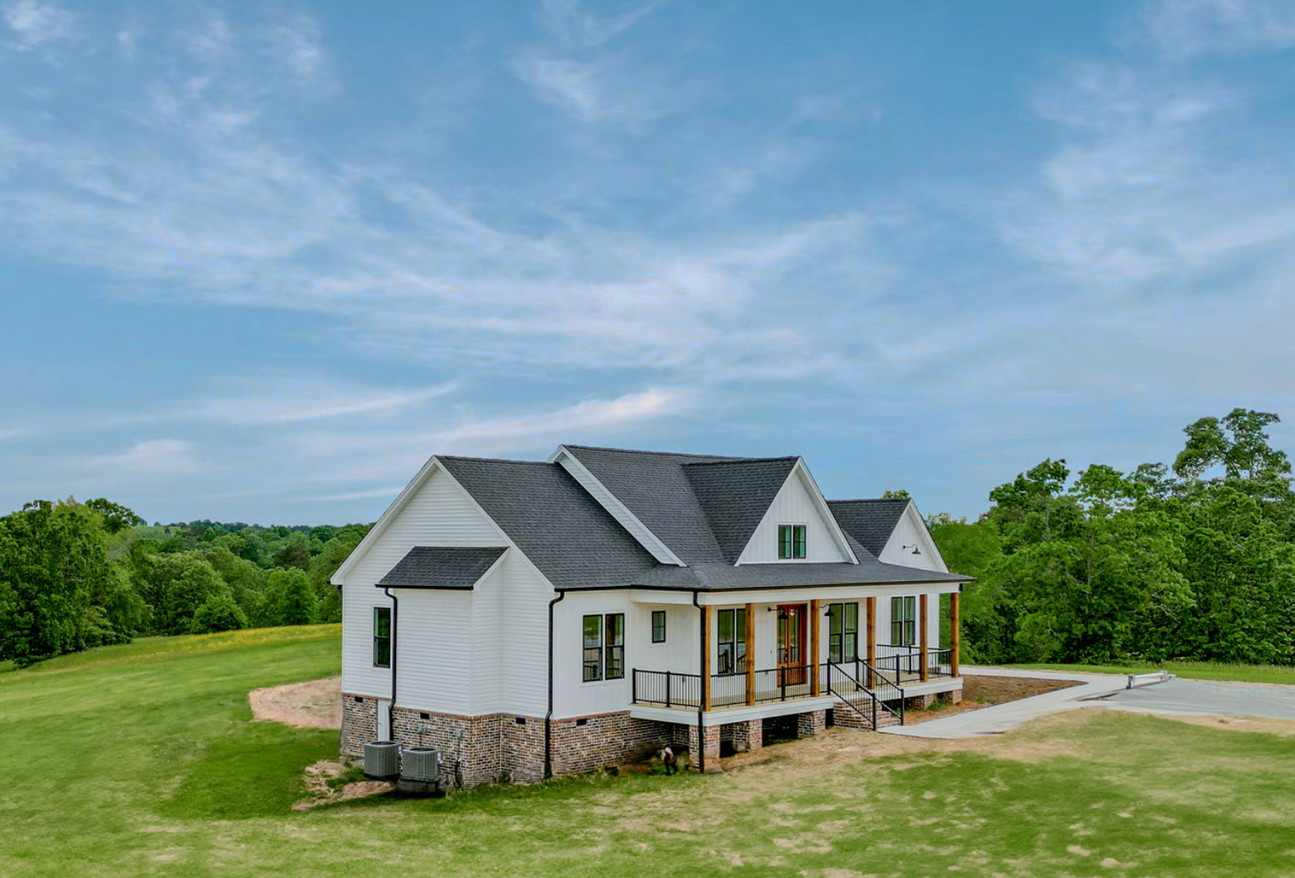 White farmhouse on a grassy lot under a blue sky with trees in the background