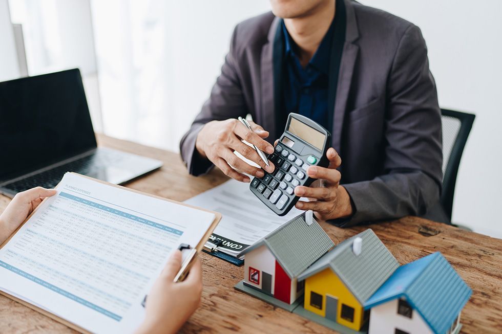 Person using a calculator beside a clipboard, laptop, and model house on a desk