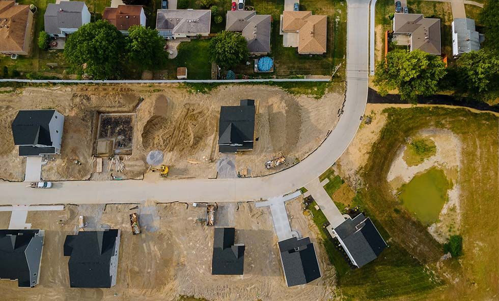 Aerial view of a suburban street with houses, construction lots, and a grassy vacant area.