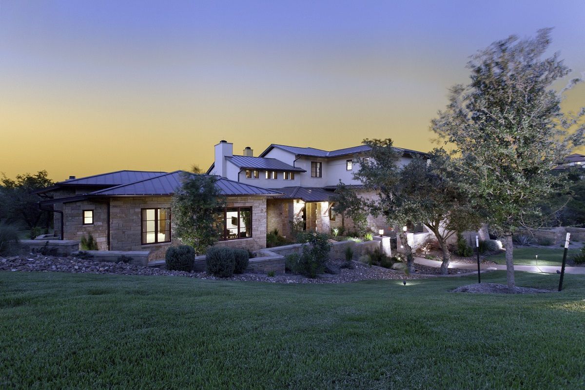 Stone house at dusk, lit windows, lush landscaping and a green lawn.
