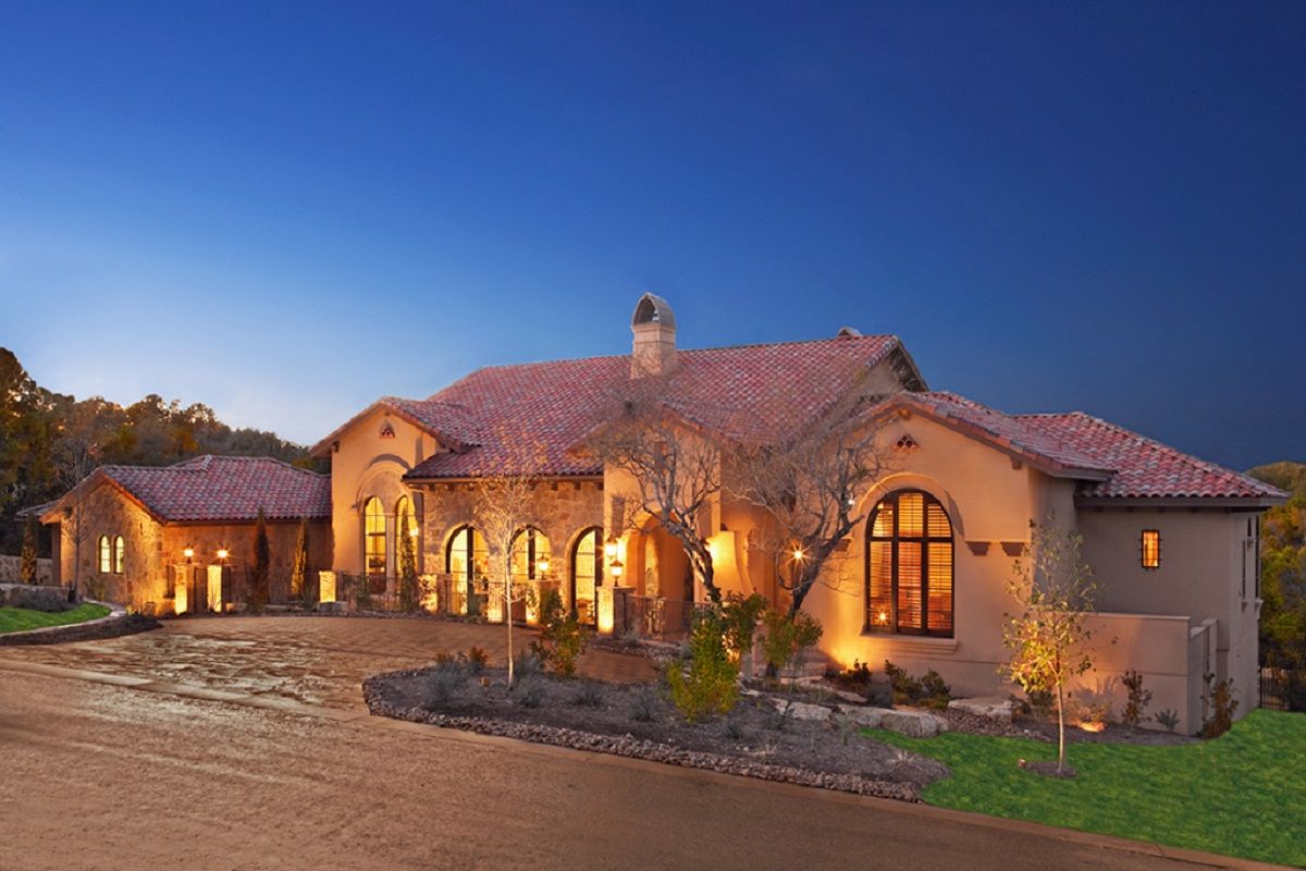 Luxurious beige stucco house with red-tiled roof at dusk, illuminated by exterior lights.