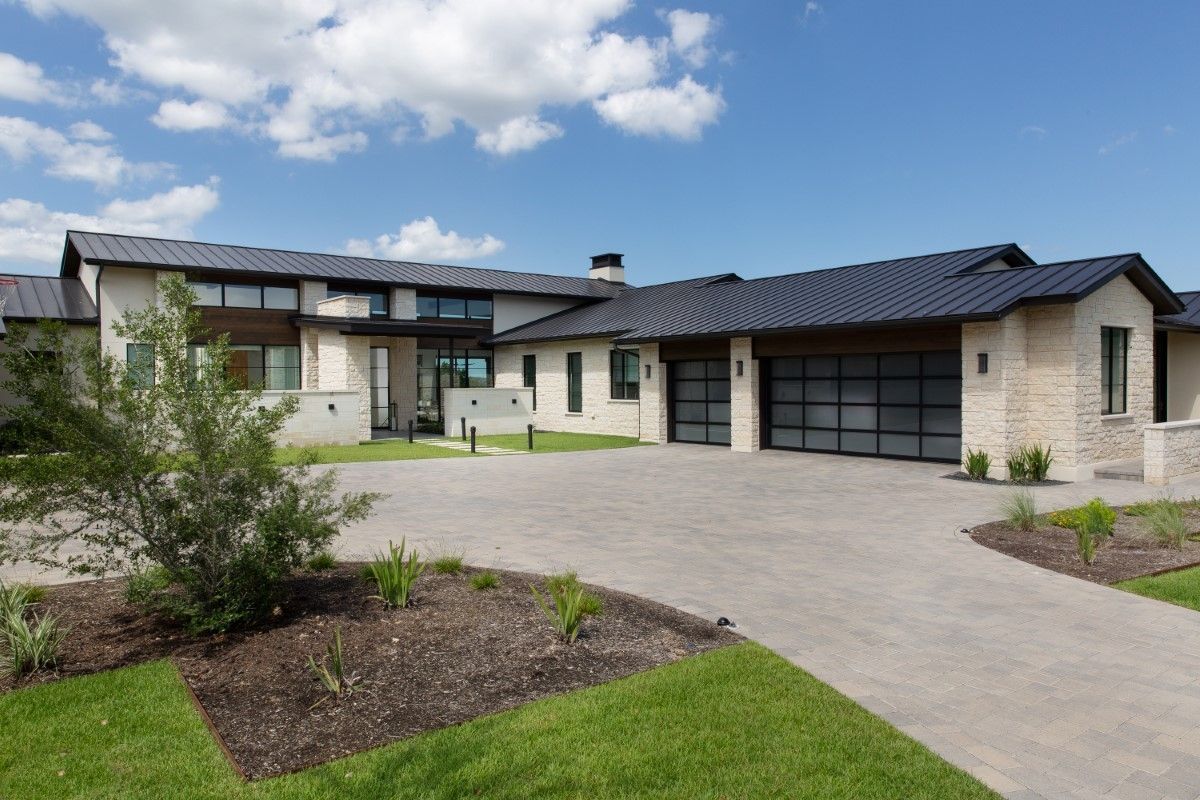 Modern house with light-colored stone exterior, dark roof, and black garage doors, set in a sunny landscape.