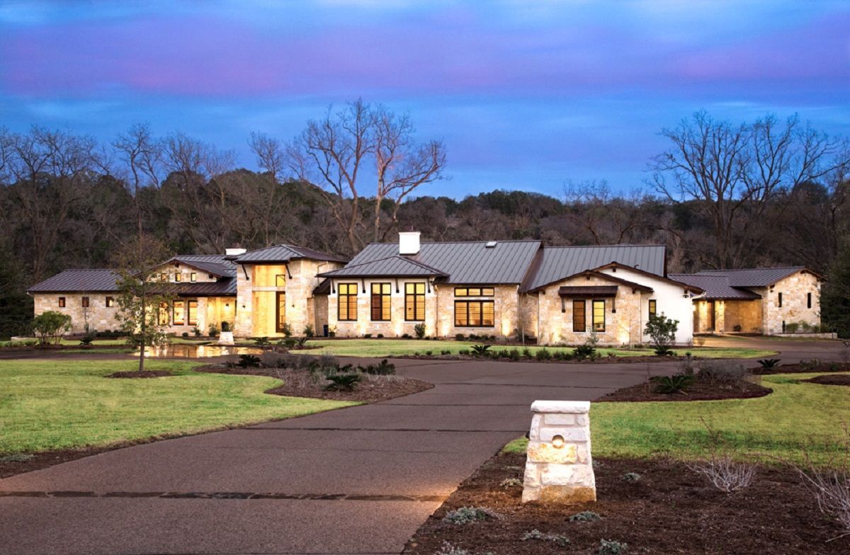 Large, stone house with a long driveway, set against a twilight sky.