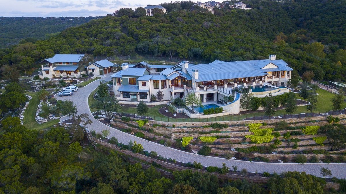 Aerial view of a large, luxurious house with a blue roof, pool, and driveway on a hillside, surrounded by trees.
