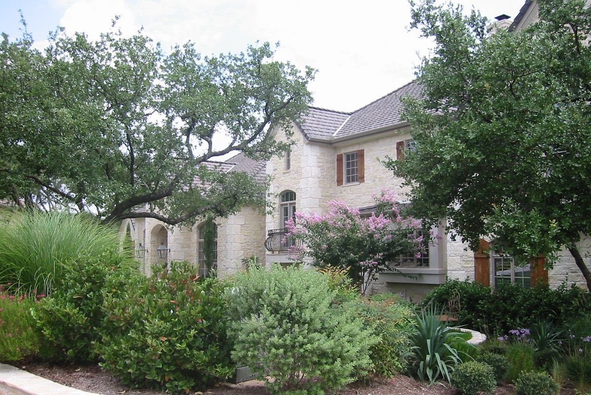 Stone house with lush landscaping; trees, bushes, and flowers in front.