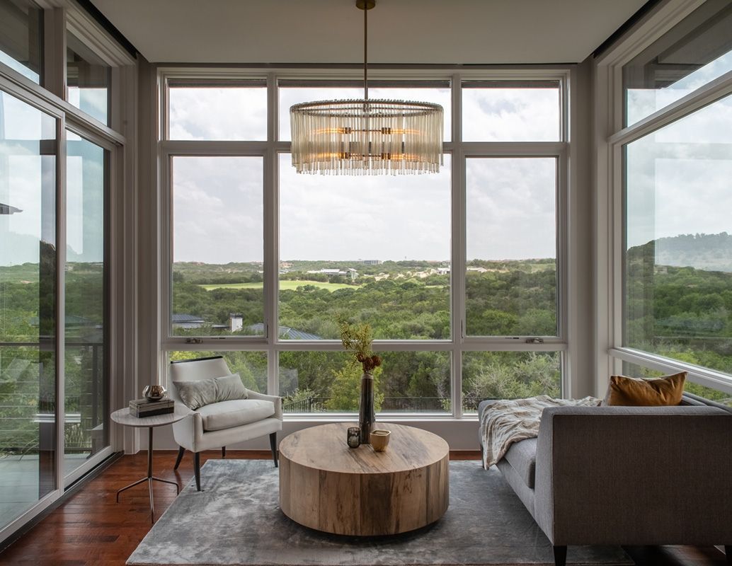 Living room with large windows overlooking a green landscape. Includes furniture, a chandelier, and a rug.