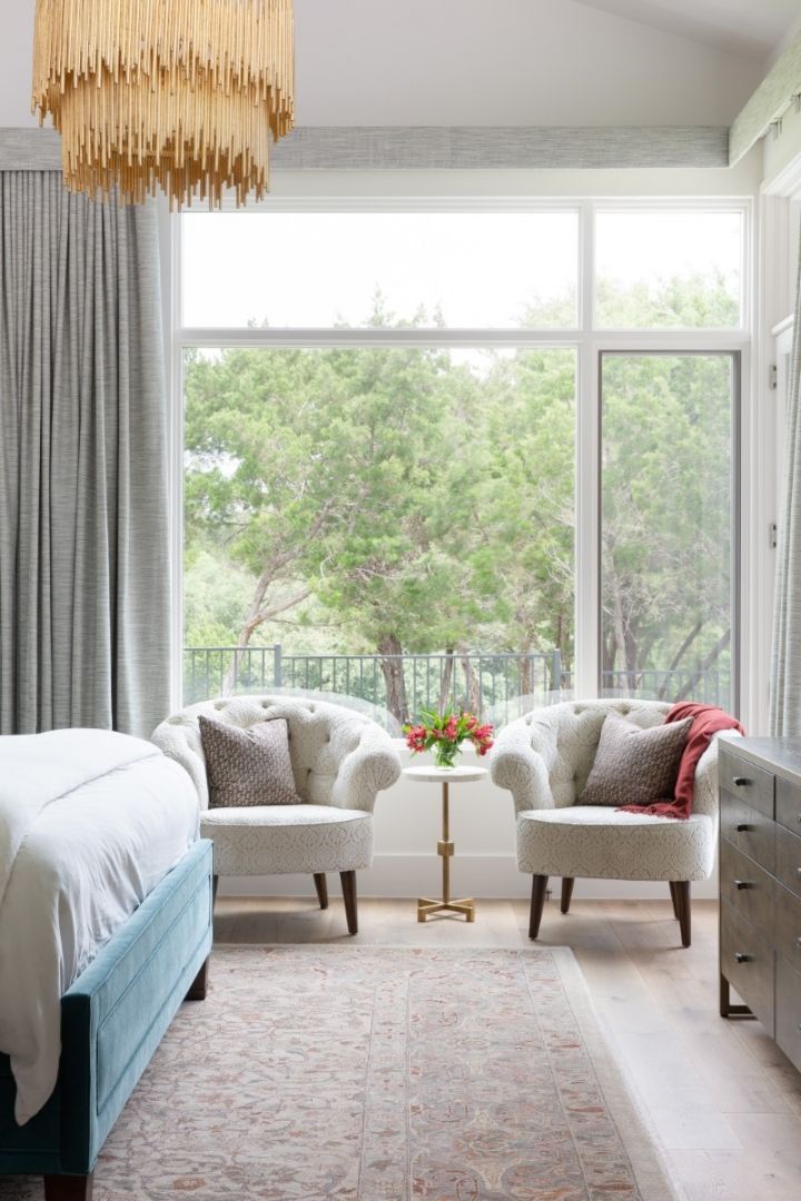Bedroom with two armchairs facing a large window, trees visible outside. Teal bed, chandelier, gray curtains.