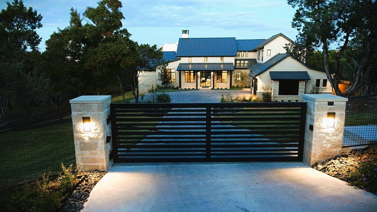 Black metal gate and stone pillars with lights illuminate a long driveway leading to a large home.
